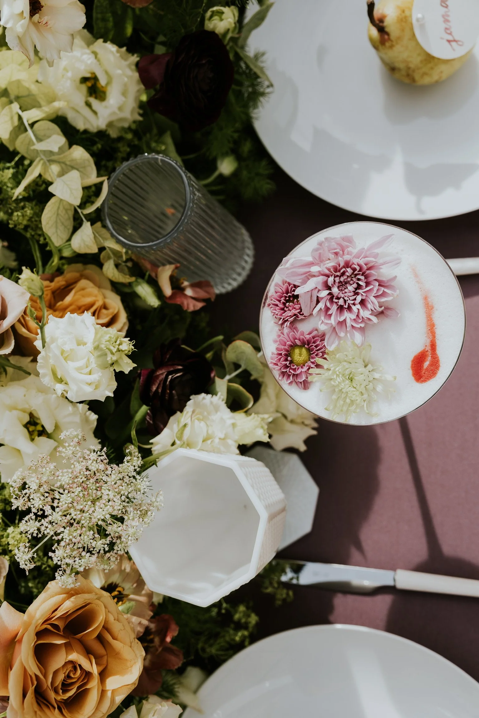 Decorative floral arrangement with white, cream, and dark red flowers, alongside a glass of drink with pink flowers and orange syrup, on a table with a white plate.
