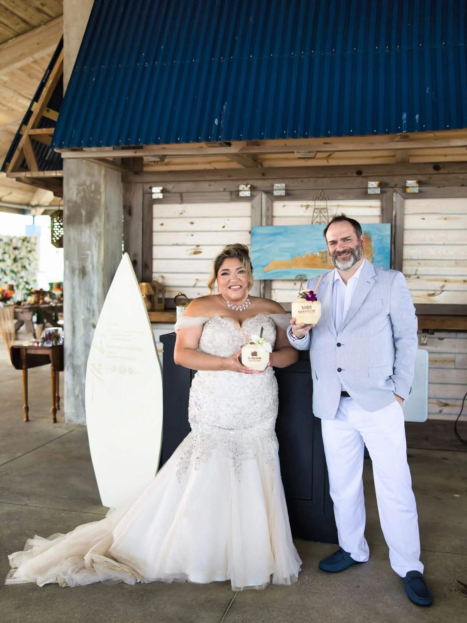 A bride and groom standing together at a wedding reception, holding coconut drinks with flowers. The bride wears a strapless white lace wedding gown, and the groom is dressed in a light blue blazer with white pants. They are smiling and posing for the camera inside a rustic venue with wooden walls and a painted landscape picture in the background.