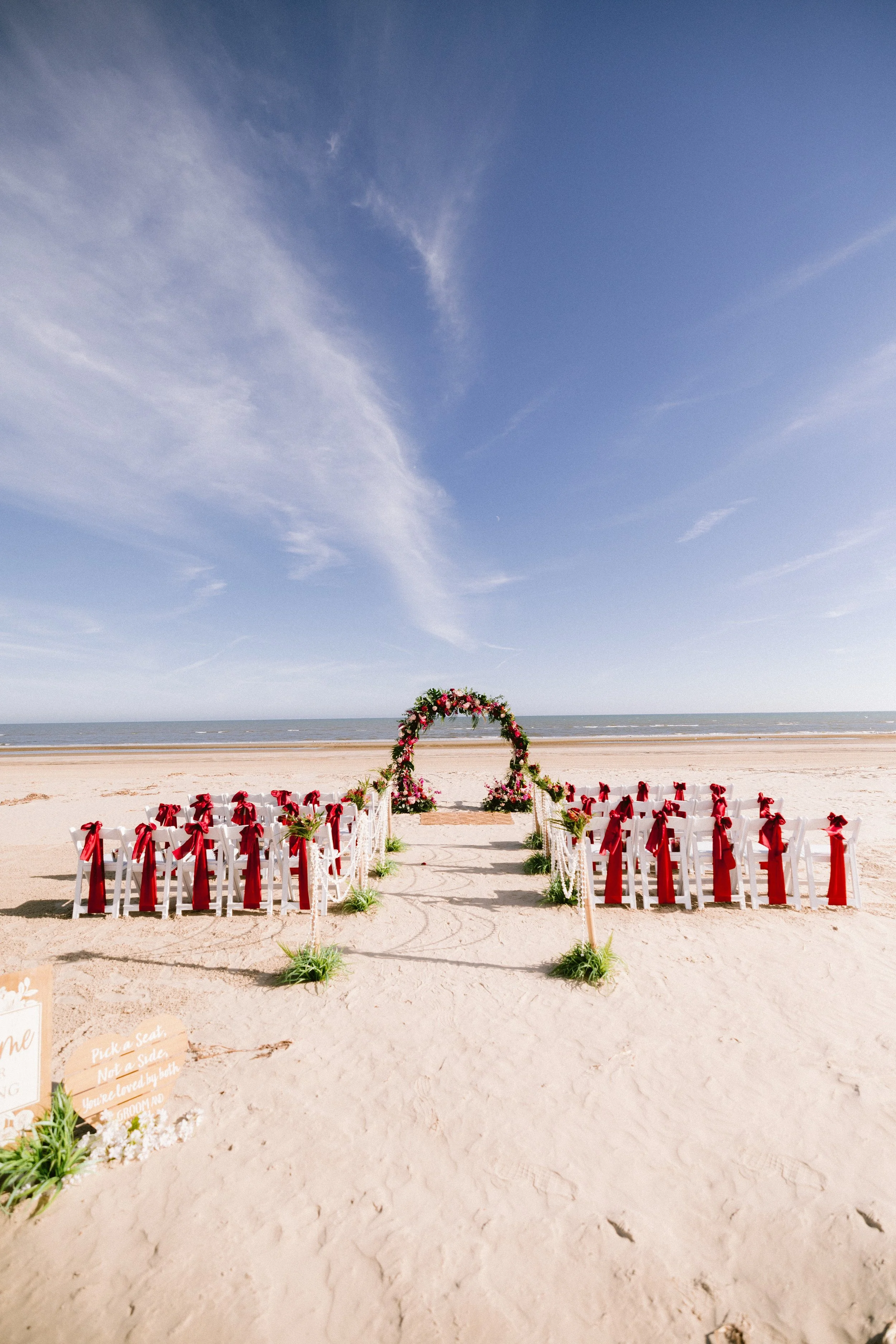 Empty beach wedding setup with white chairs adorned with red ribbons facing a floral arch over the aisle, on a sandy shore under a blue sky.