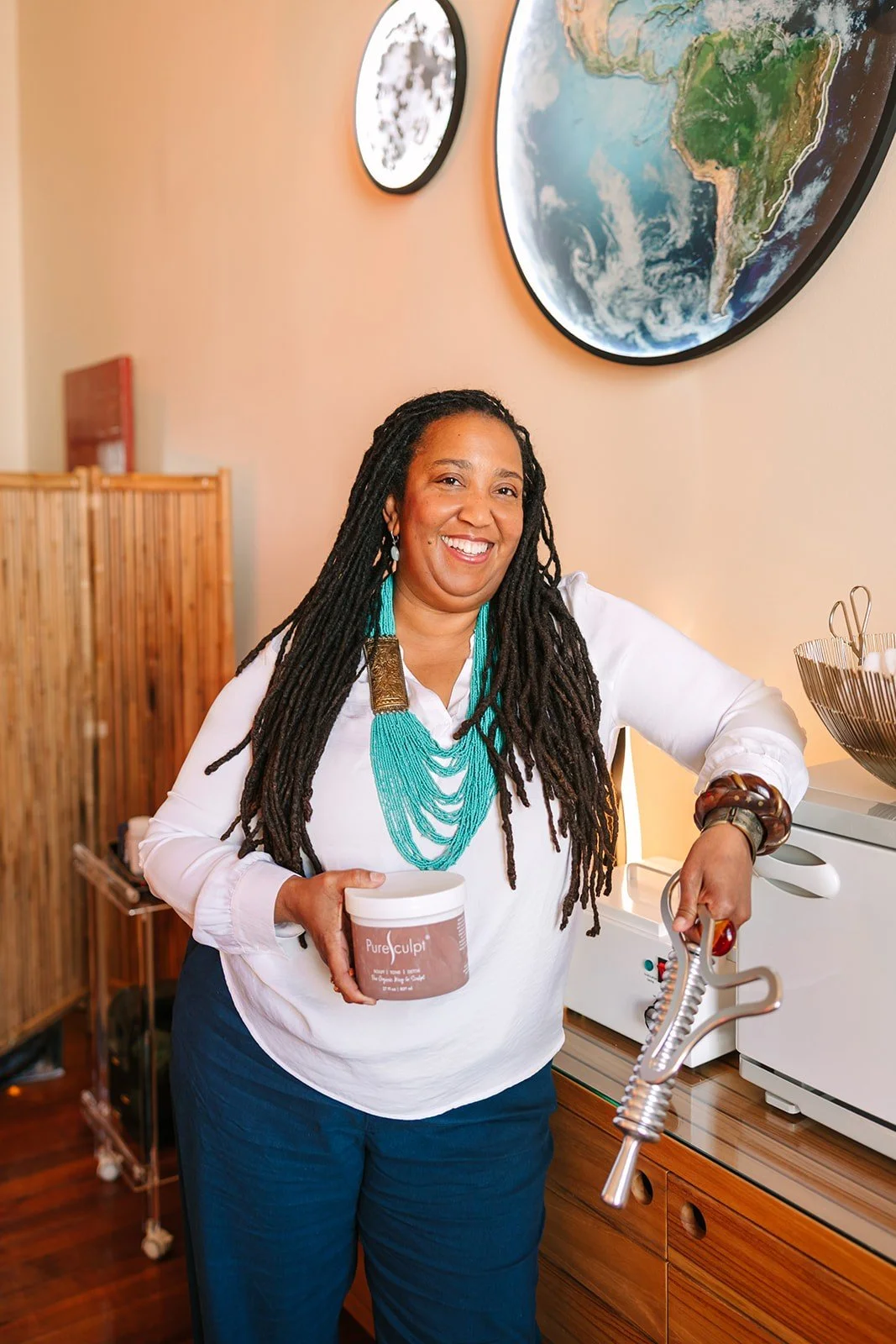 A woman with dreadlocks and a white blouse smiling and holding a container labeled 'Pure Sculpt' in one hand and a kitchen tool in the other, standing in a room with a map of Earth on the wall.