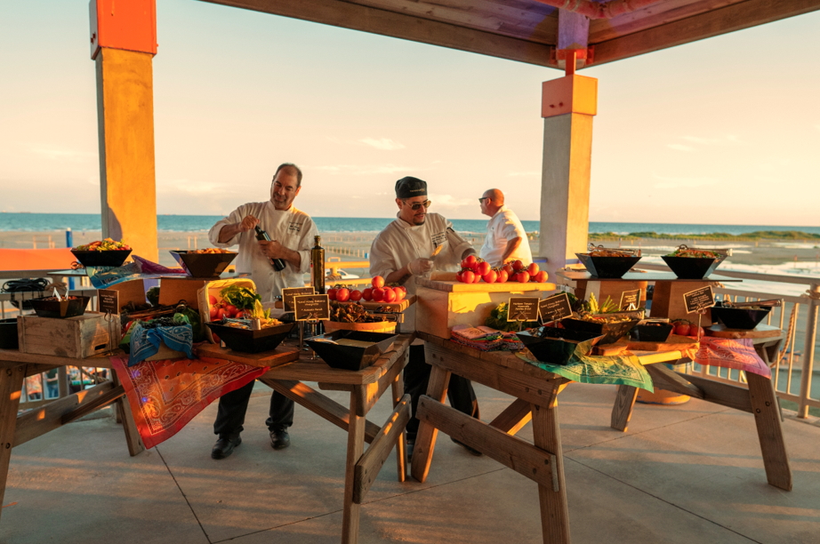 Chefs preparing and serving food at an outdoor event on a beachside patio during sunset.