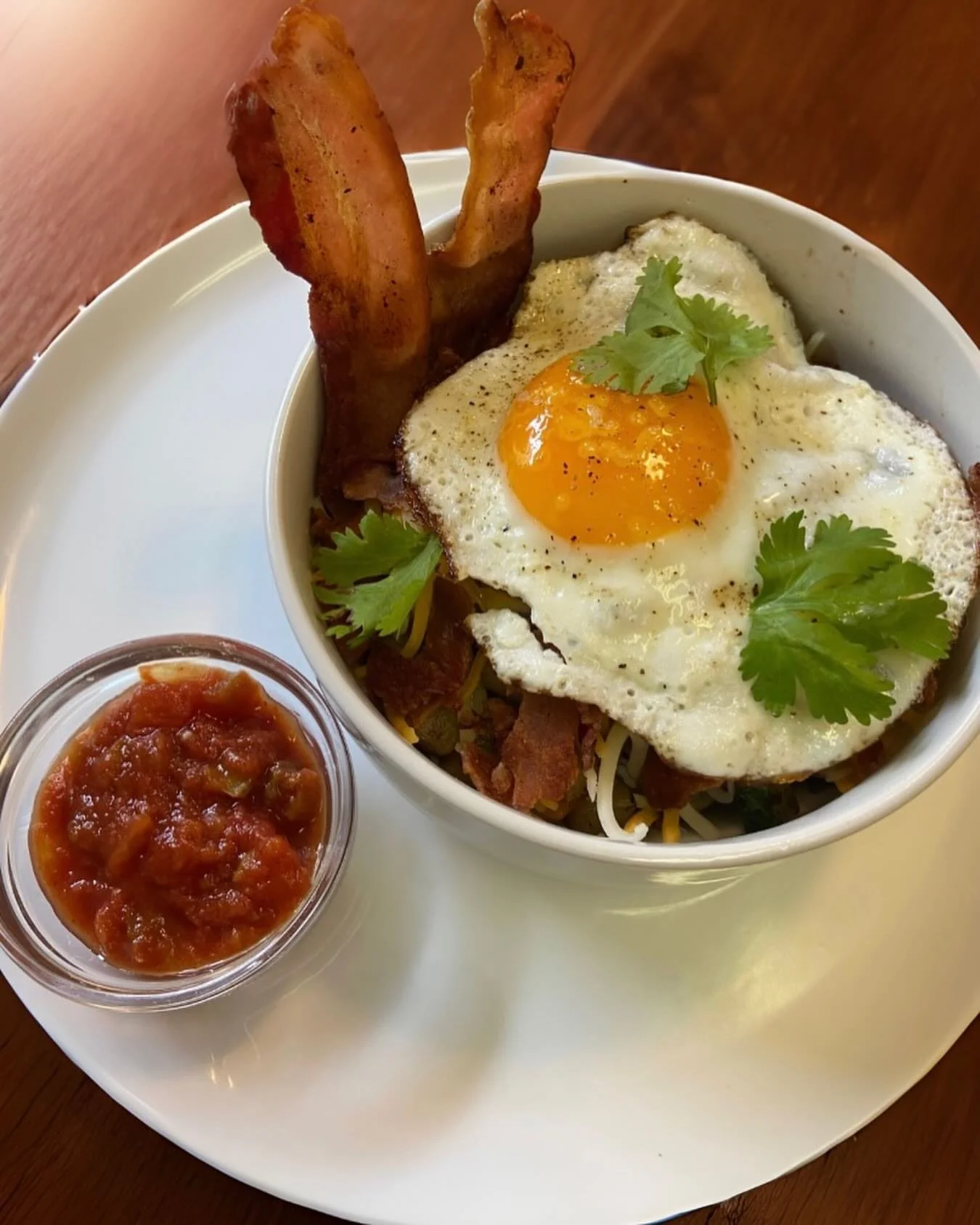 A bowl of food with a fried egg, bacon, cilantro, and other toppings. A side of salsa in a small container is on a white plate.