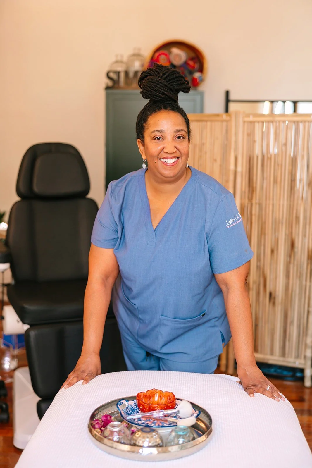 A woman in blue scrubs standing behind a table with a decorative tray of assorted items, smiling at the camera in a room with a black chair, a green cabinet, and a wooden room divider.
