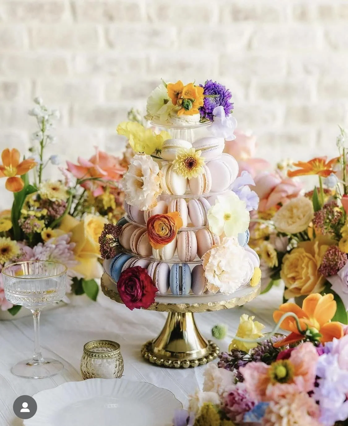 A dessert display with a tall tower of pastel-colored macarons decorated with flowers, surrounded by a floral centerpiece on a decorated table.