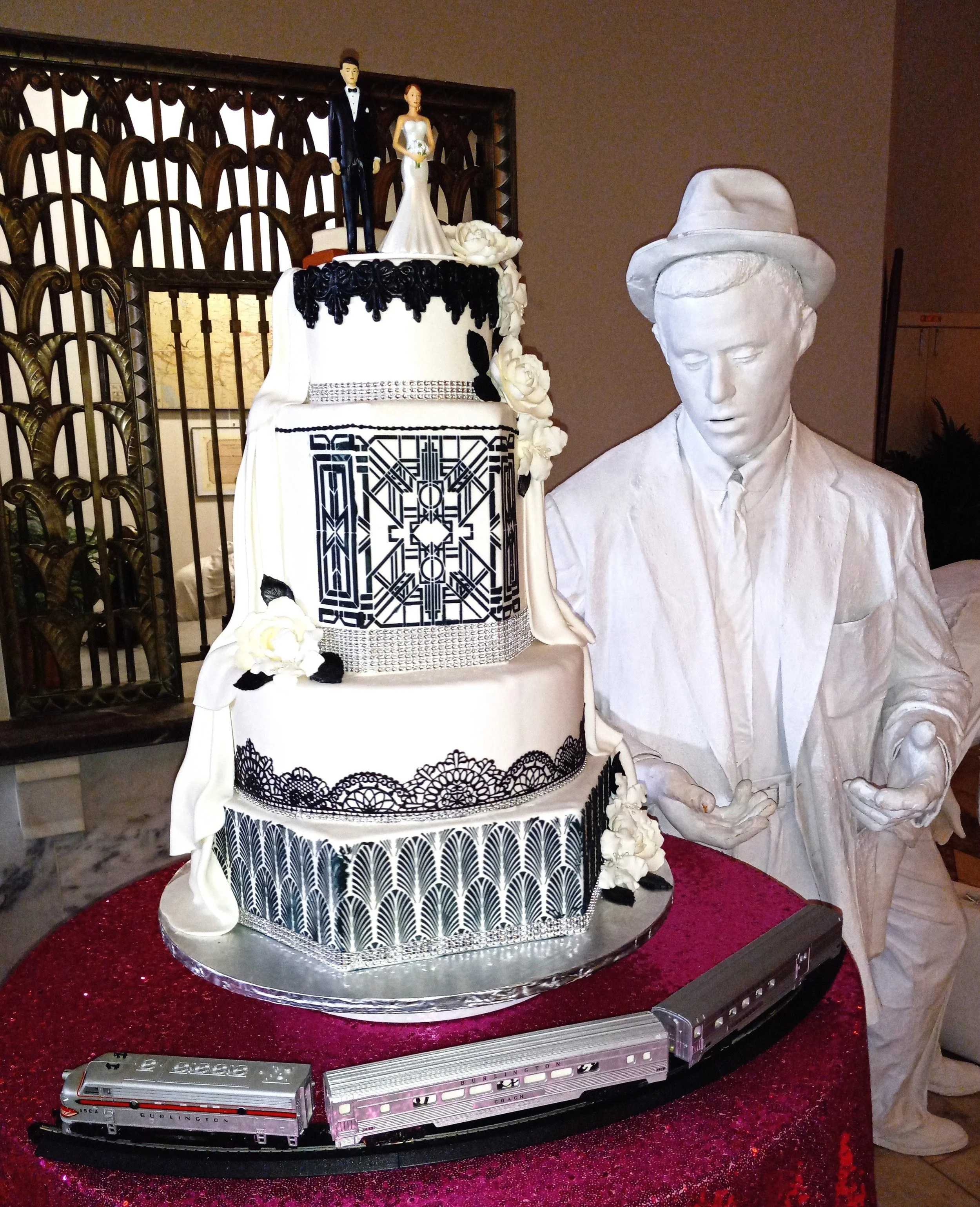 A two-tier wedding cake decorated with pink and white roses, blue hydrangeas, and greenery on a wooden slab. The cake topper reads 'Mrs & Mrs' and is surrounded by hanging fairy lights. The cake is on a sparkling gold tablecloth, flanked by two tall champagne flutes, with a brick wall and ornate candleholders reflected in a mirror behind.