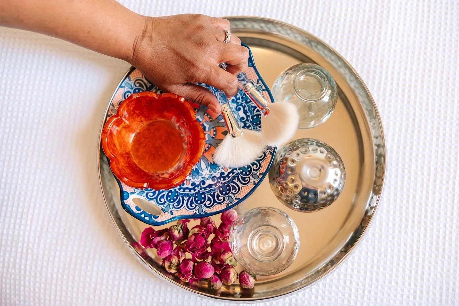 A decorative silver tray holding various items including a colorful tray with a floral pattern, a red glass bowl, two small clear glass bottles, a silver decorative orb, and a bunch of dried pink rosebuds. A hand with a ring is holding a face brush over the items.