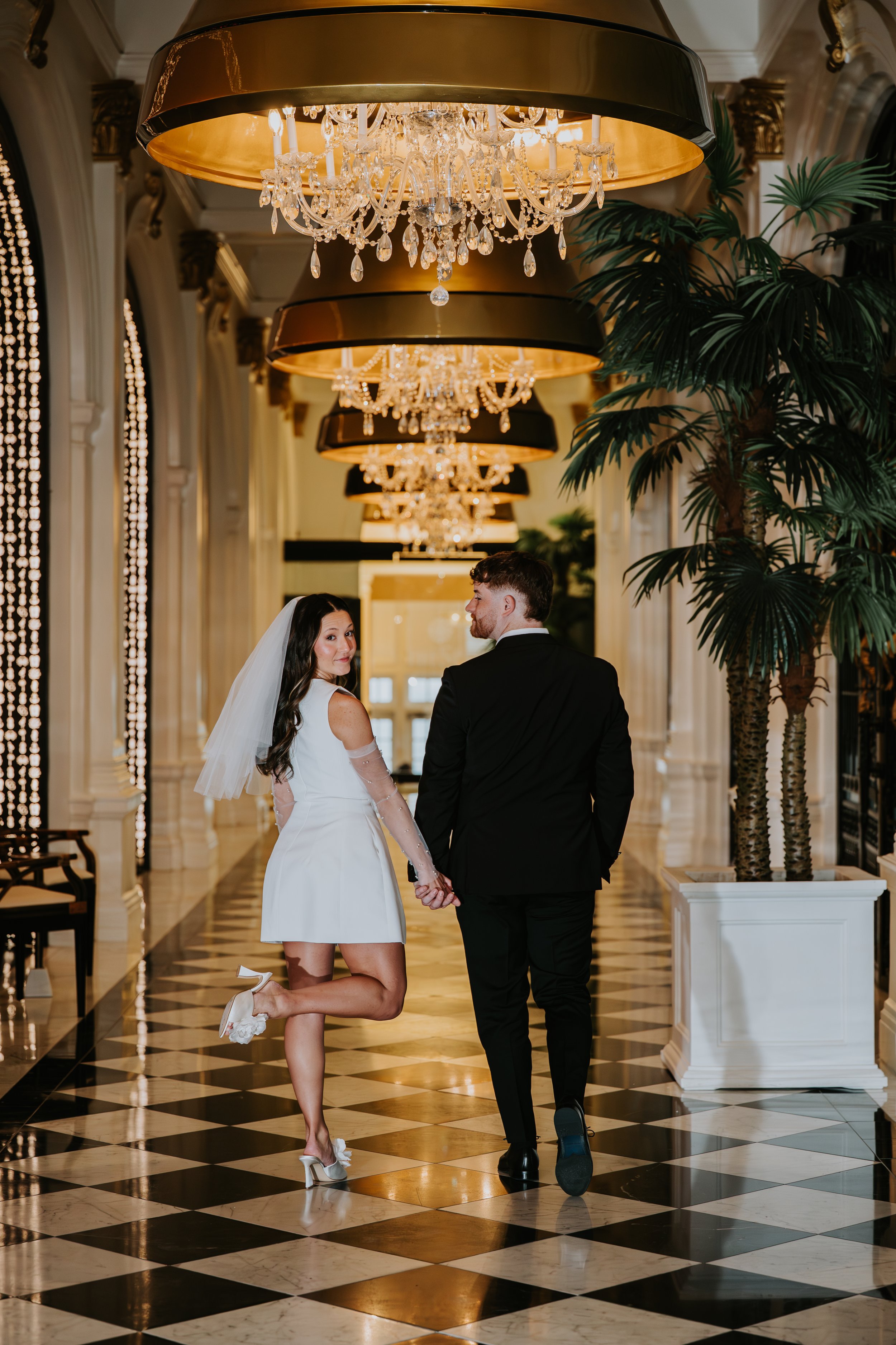 A bride and groom holding hands in an elegant hotel lobby with chandeliers, large potted plants, and checkered marble floors. The bride is kneeling on one knee, wearing a white dress and veil, while the groom is standing and looking at her.