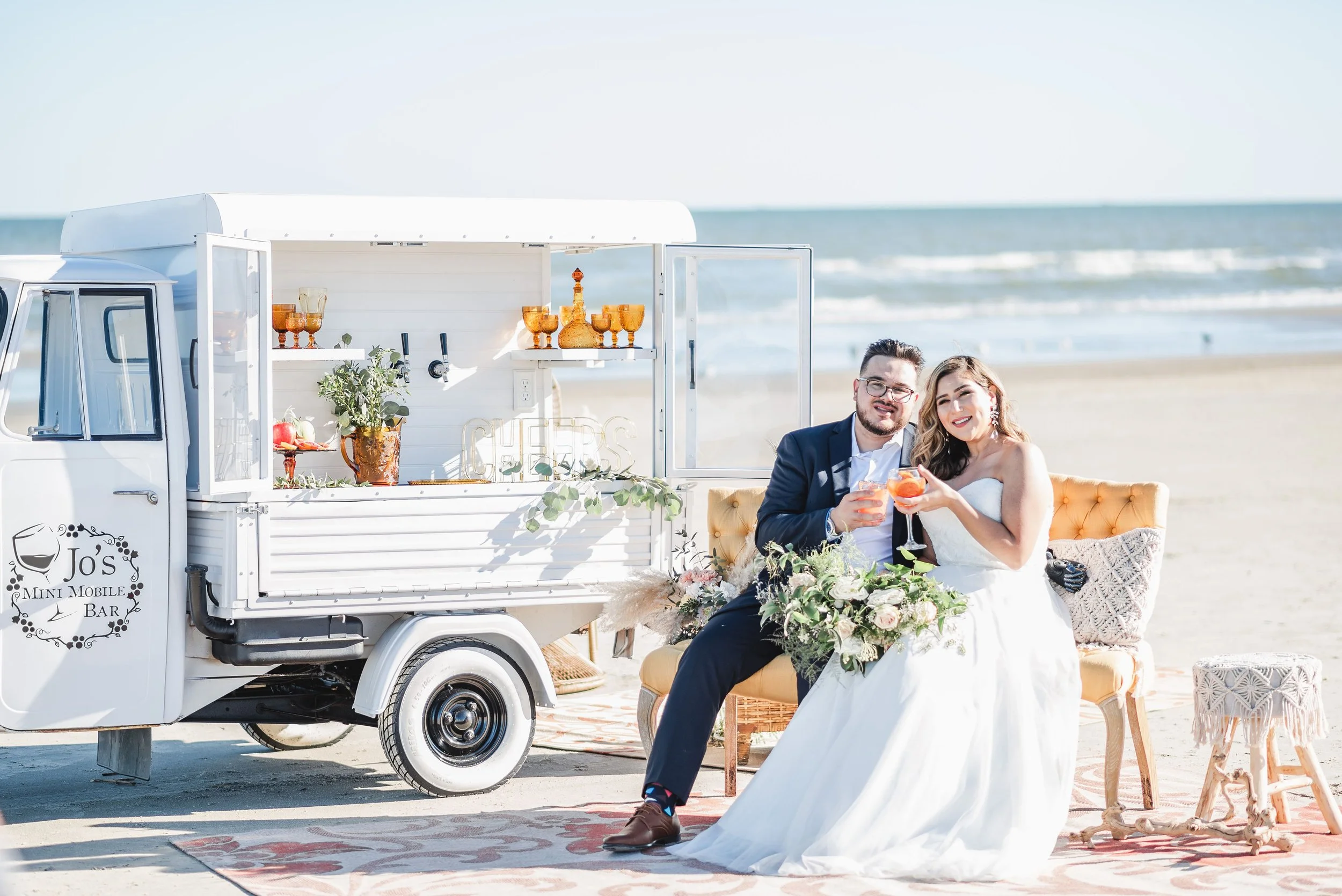 A bride and groom sitting on a yellow sofa at a beach, holding drinks and smiling, with a white mini mobile bar cart nearby.