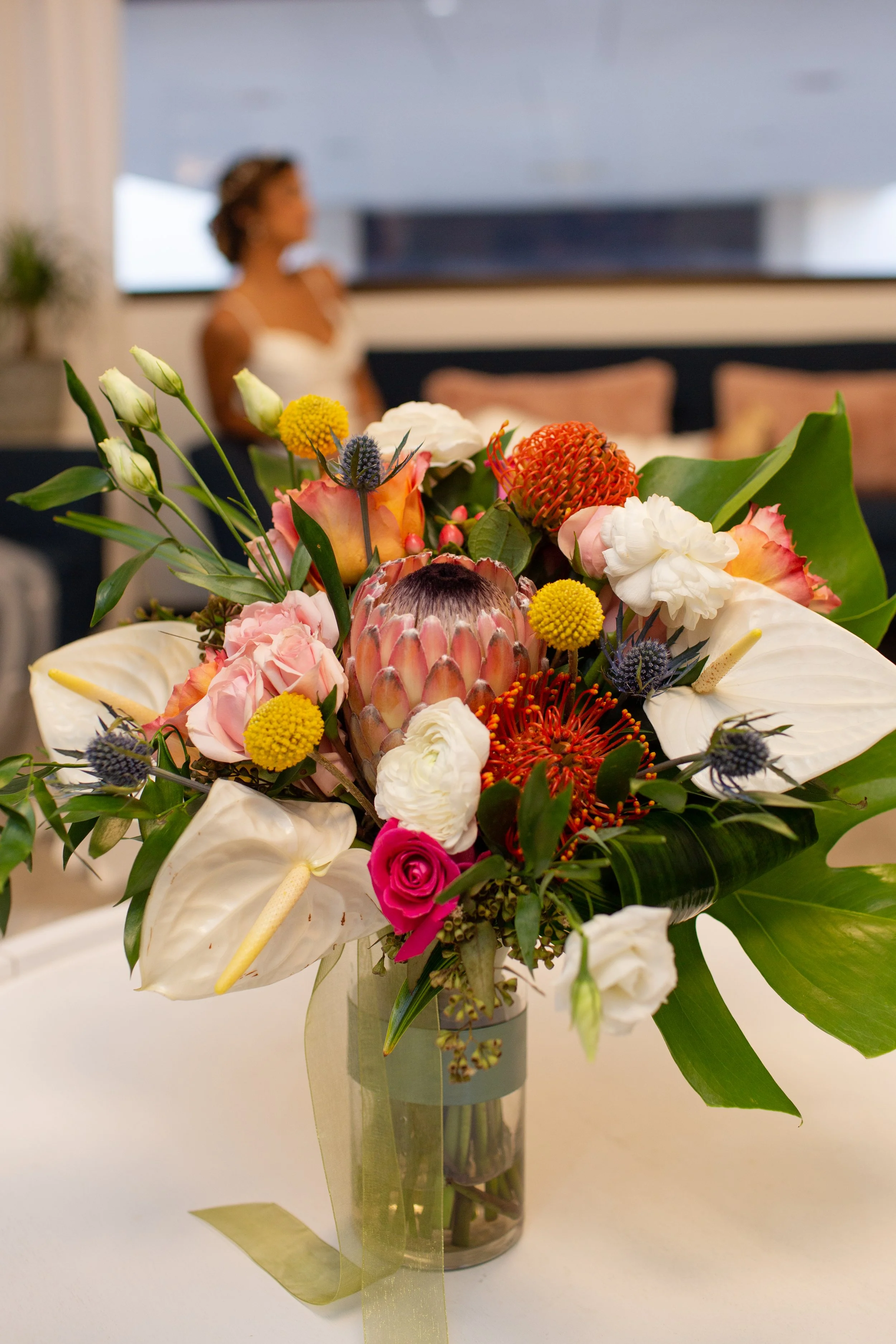A colorful flower bouquet in a glass vase, featuring white lilies, pink roses, yellow billy balls, orange protea, and other assorted flowers, with a blurred woman in white dress in the background.