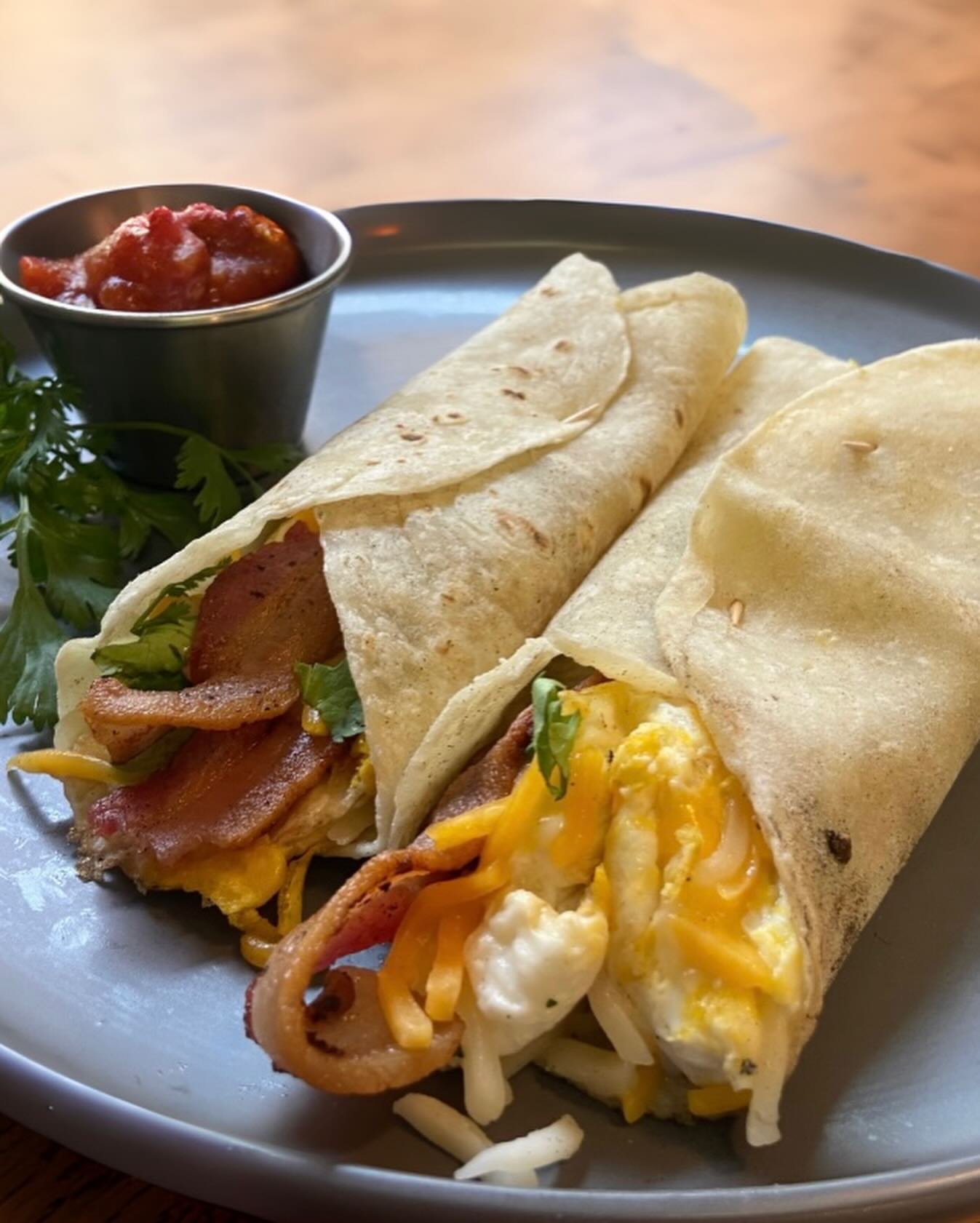 A plate holding two breakfast burritos with a small cup of salsa on the side, garnished with cilantro.