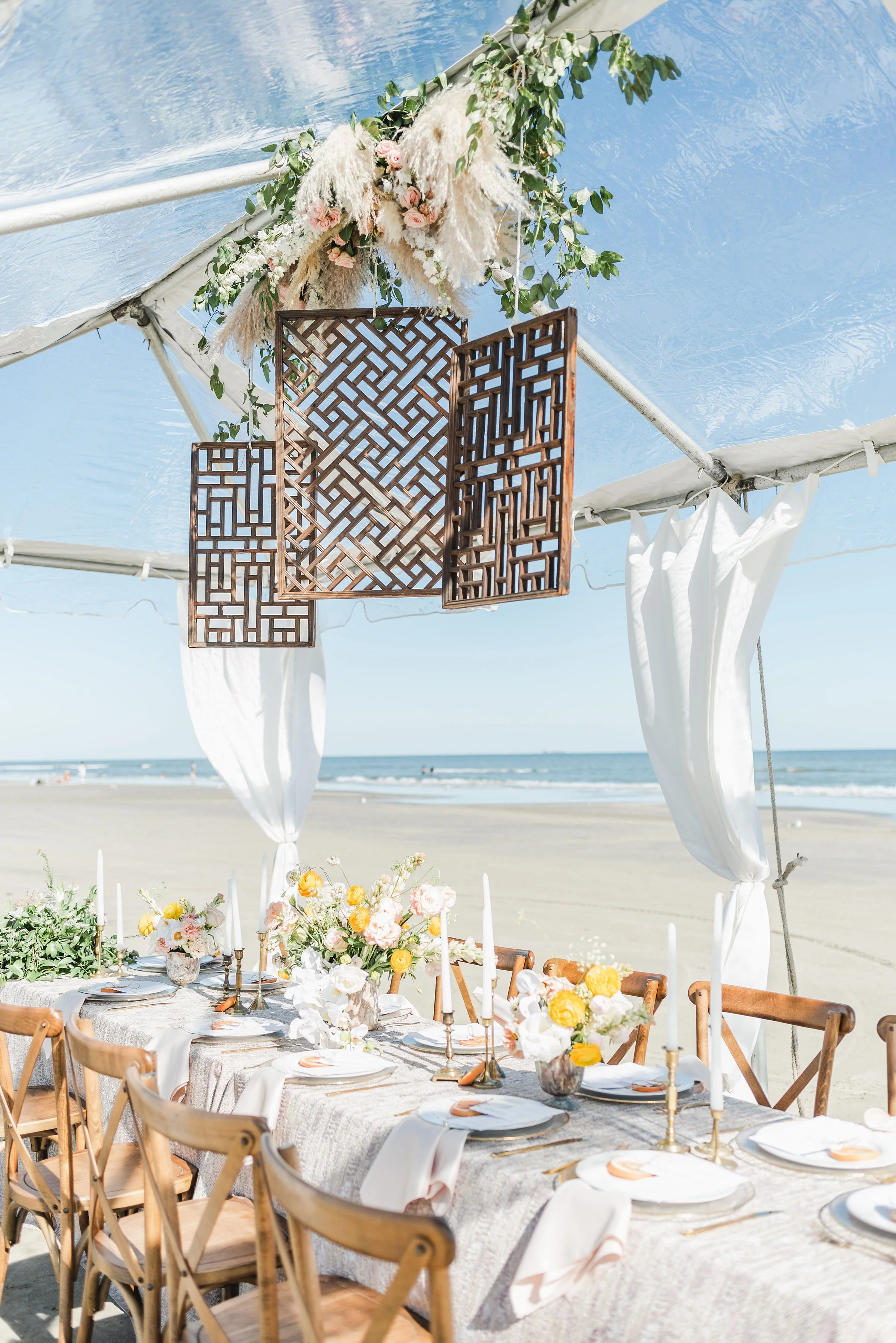 Beachside wedding or event setup with a long dining table decorated with flowers and candles under a canopy with hanging wooden panels and floral arrangements.