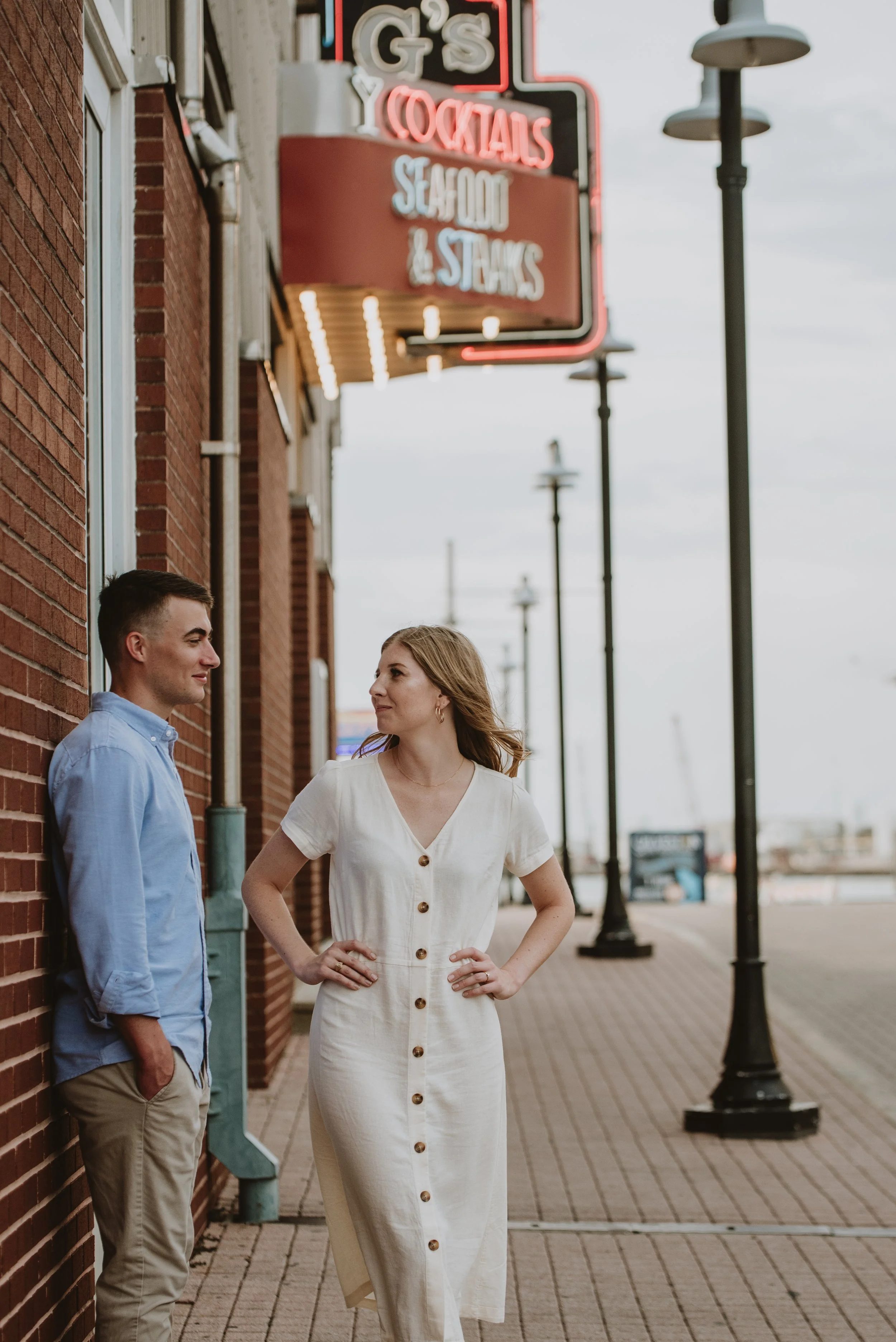 A man and woman standing and facing each other on a sidewalk outside a brick building with a neon sign that reads 'G's Cocktails Seafood & Steaks'. The woman has light brown hair and is wearing a white dress with buttons down the front, and the man has short dark hair and is wearing a light blue shirt and beige pants. Streetlights and a cloudy sky are visible in the background.