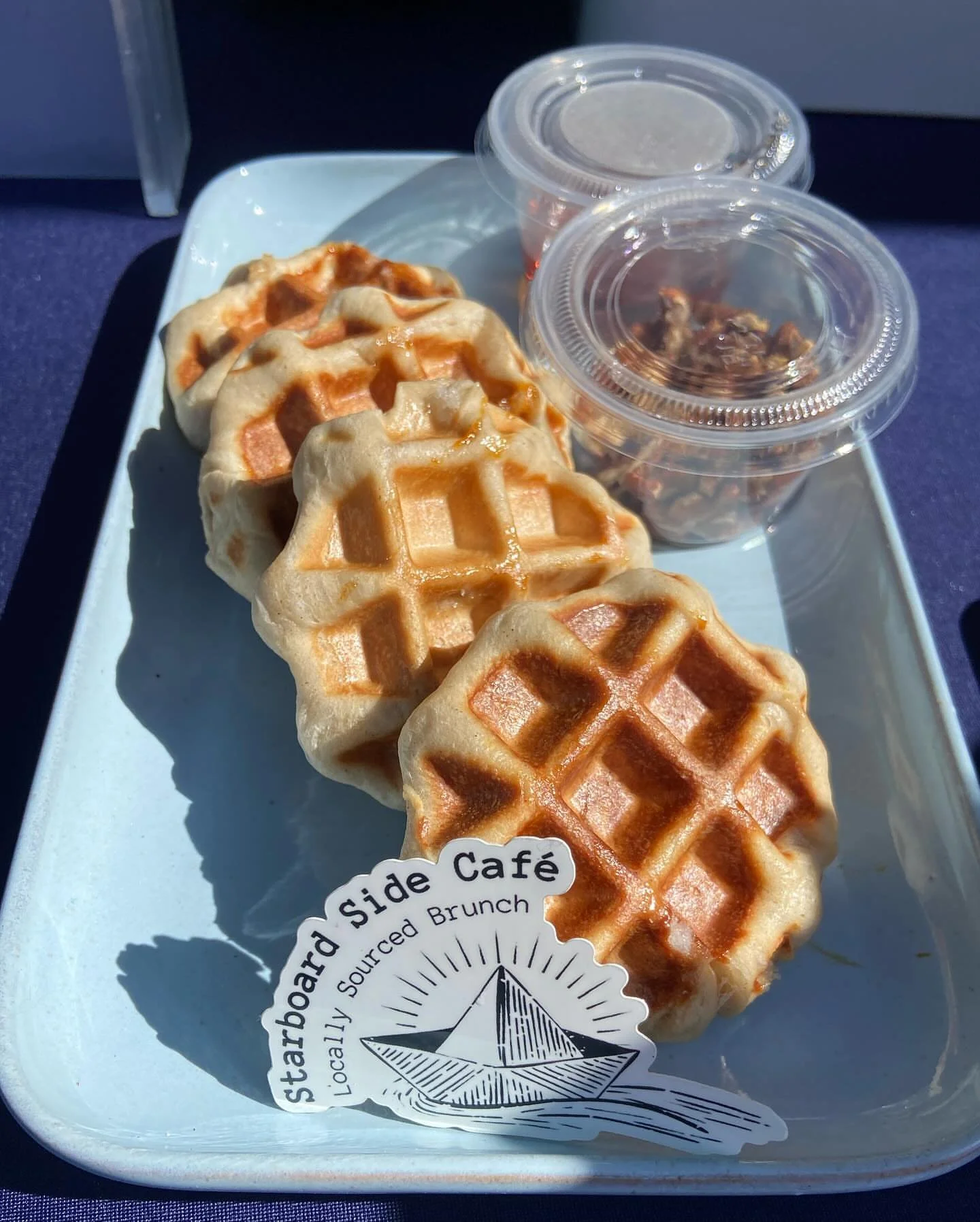 Waffles with a crispy golden-brown exterior on a white rectangular plate, accompanied by two small clear containers of syrup or toppings, with a sticker that reads 'Boardwalk Side Café' and 'Locally Sourced Brunch'.
