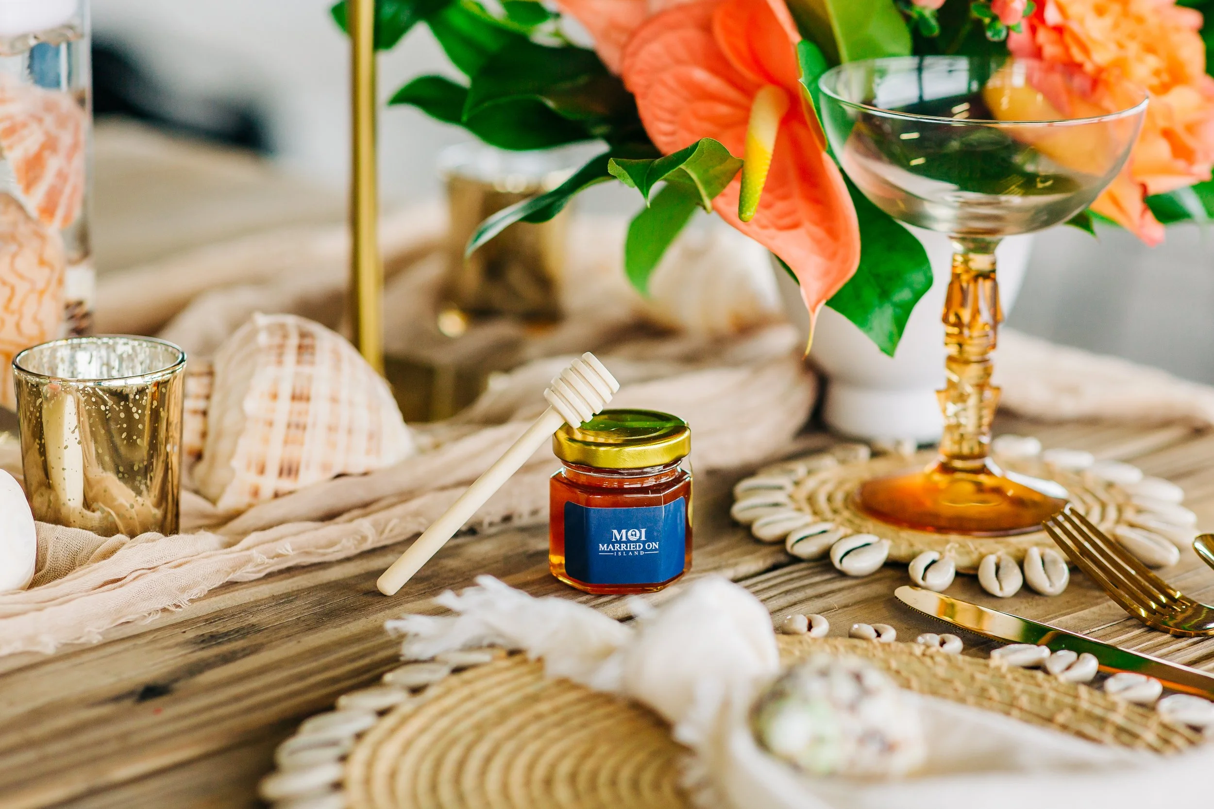 A table decorated with a bouquet of orange and pink flowers, seashells, a gold candle holder, a small jar of honey, a gold fork, and a gold spoon, creating a festive table setting.