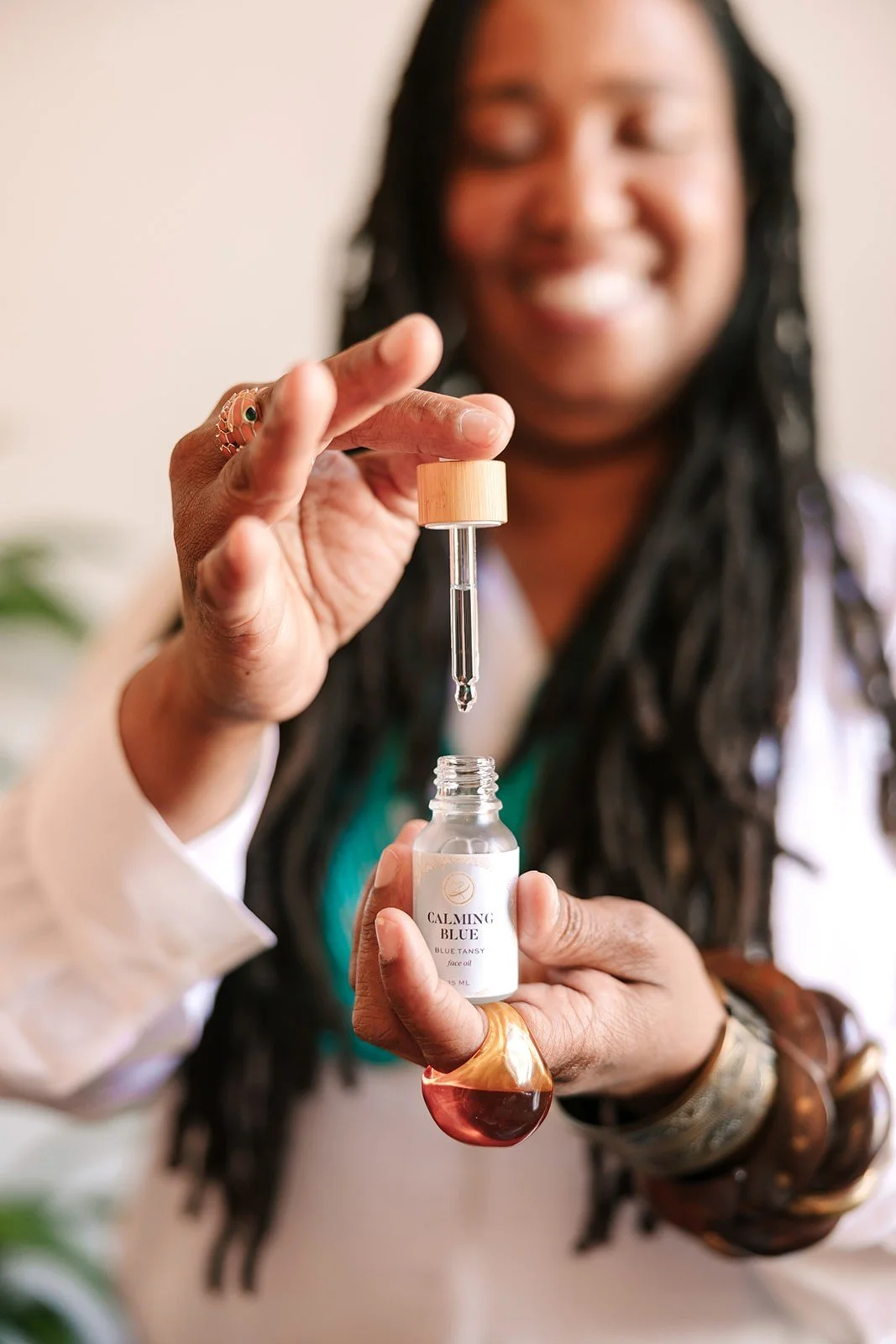 A smiling woman with long black dreadlocks holding a small dropper bottle labeled 'Calming Blue' with a liquid drop hanging from the dropper.