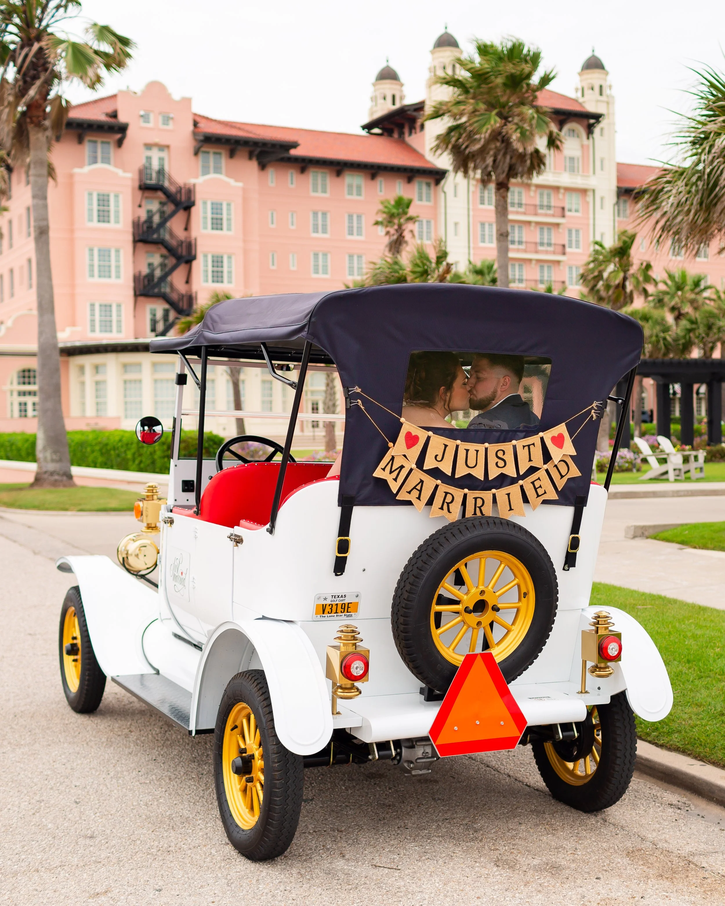 A golf cart with a couple inside, parked on a street with palm trees and a large pink building in the background. The cart has a bunting that says 'JUST MARRIED' with hearts, and a small sign on the back that says 'Texas Golf Cart'.