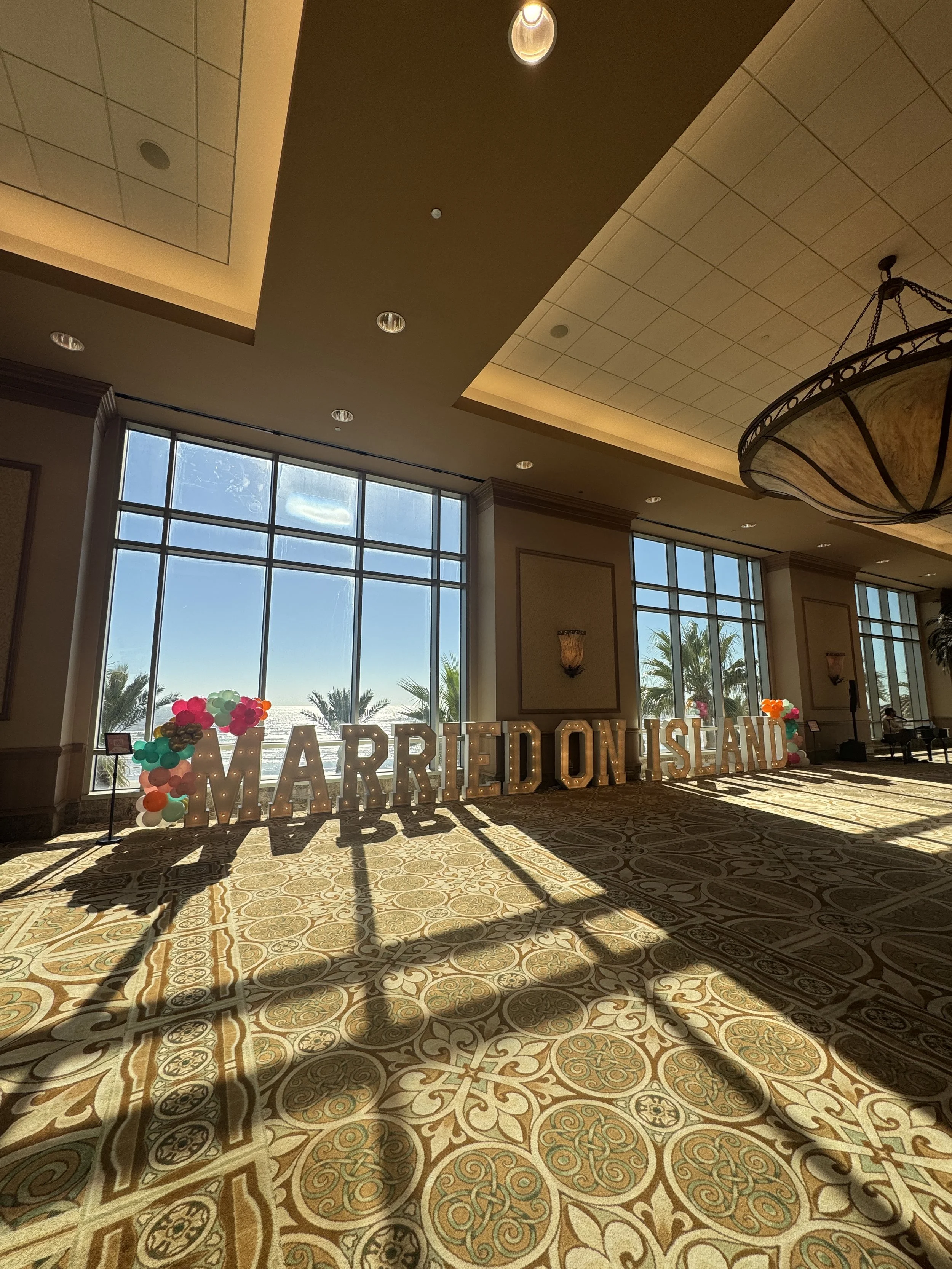 Large illuminated sign spelling 'MARRIED ON ISLAND' inside a decorated hall with large windows, balloons, and palm trees outside.