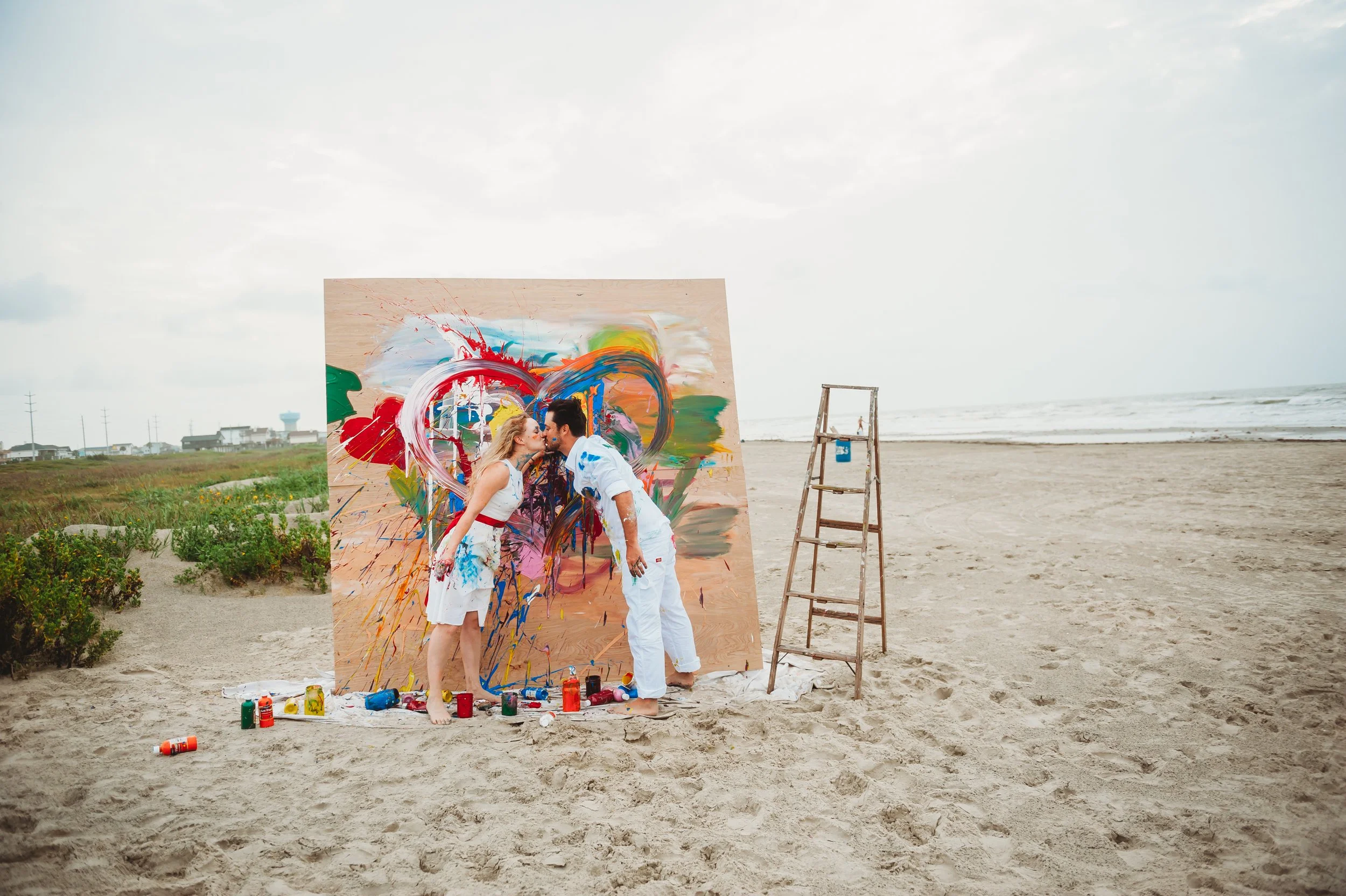 Two people kissing and smiling in front of a large, colorful abstract painting on a beach. Paint bottles and brushes are scattered on the sand, and a wooden ladder stands nearby. The sky is cloudy and the ocean is in the background.