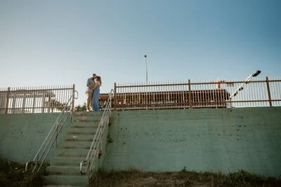 A couple standing close together on a concrete platform near a railing, overlooking a blue sky with a street lamp.