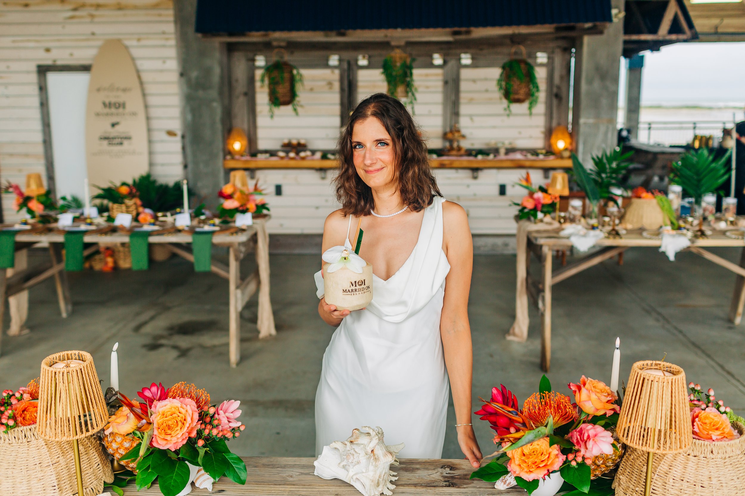 A woman in a white dress holding a tropical cocoon with a flower on top, standing in front of a decorated table with flowers and lamps at a beachside wedding reception.
