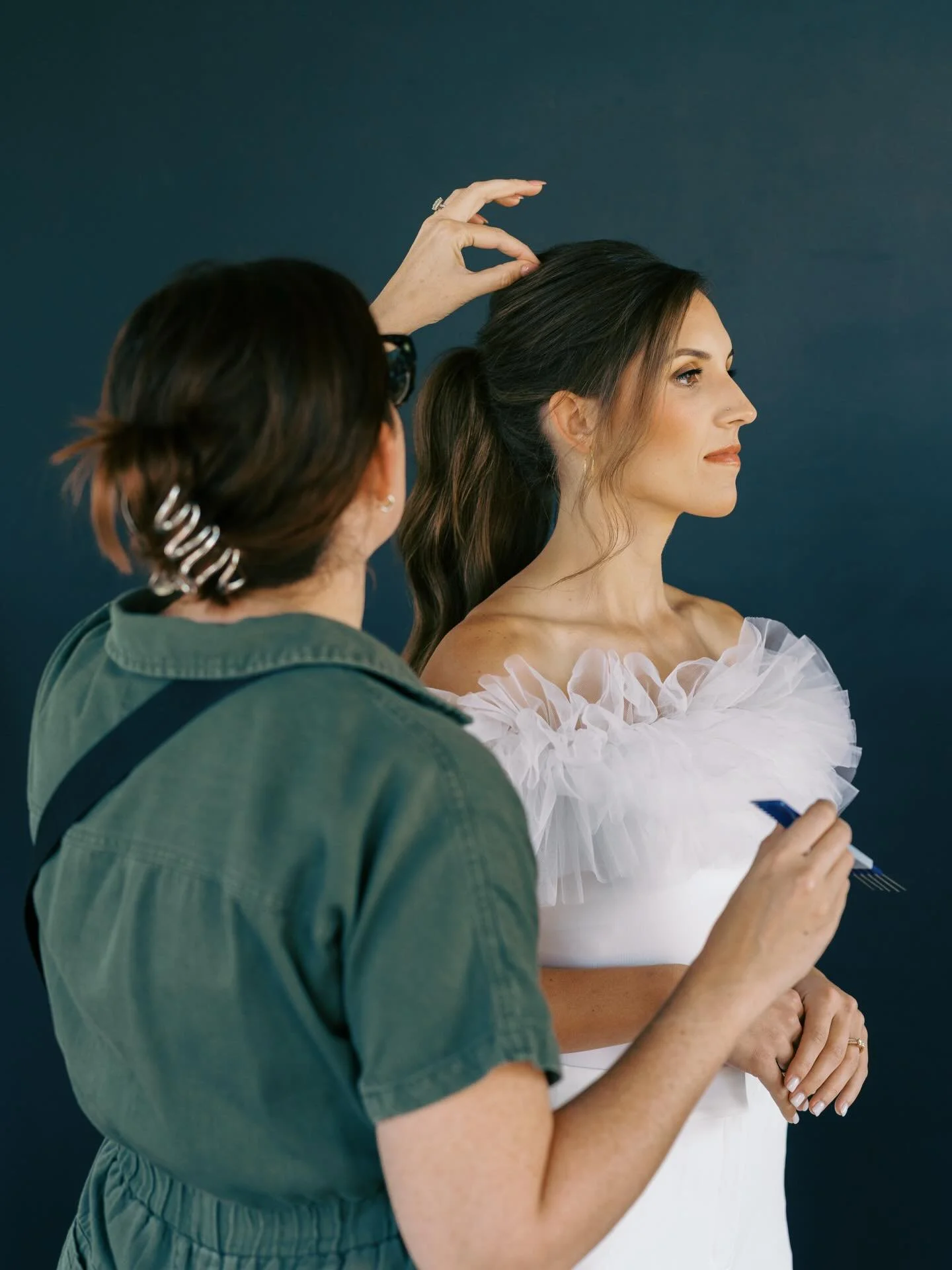 A woman with long brown hair in a ponytail, wearing an off-the-shoulder white ruffled top, holds a pen and looks to the right. A person with short dark hair and glasses, wearing a green jacket, stands beside her adjusting her hair against a dark blue background.
