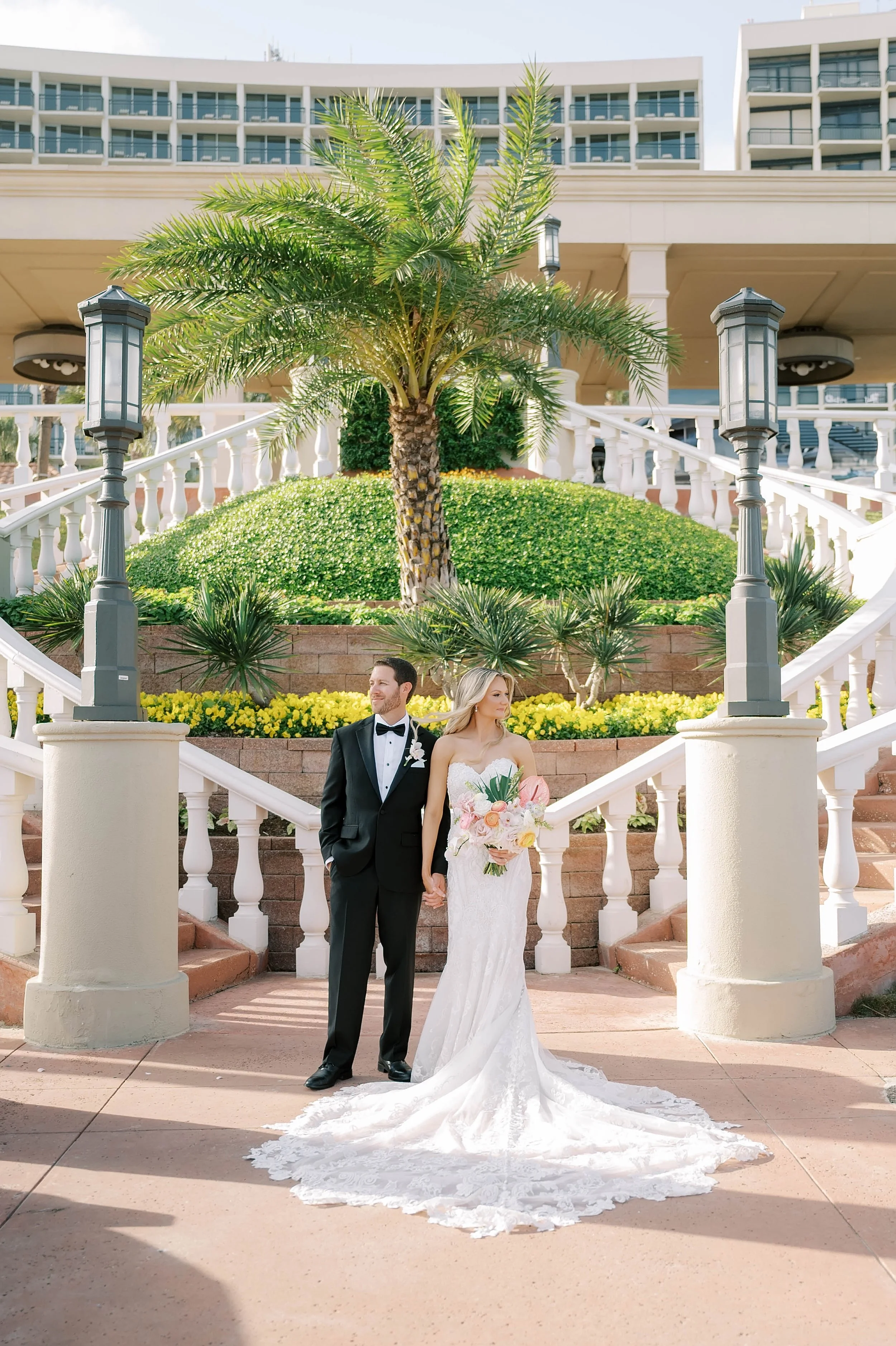 A bride and groom holding hands in front of a staircase with a palm tree on a landscaped area during their wedding photoshoot.