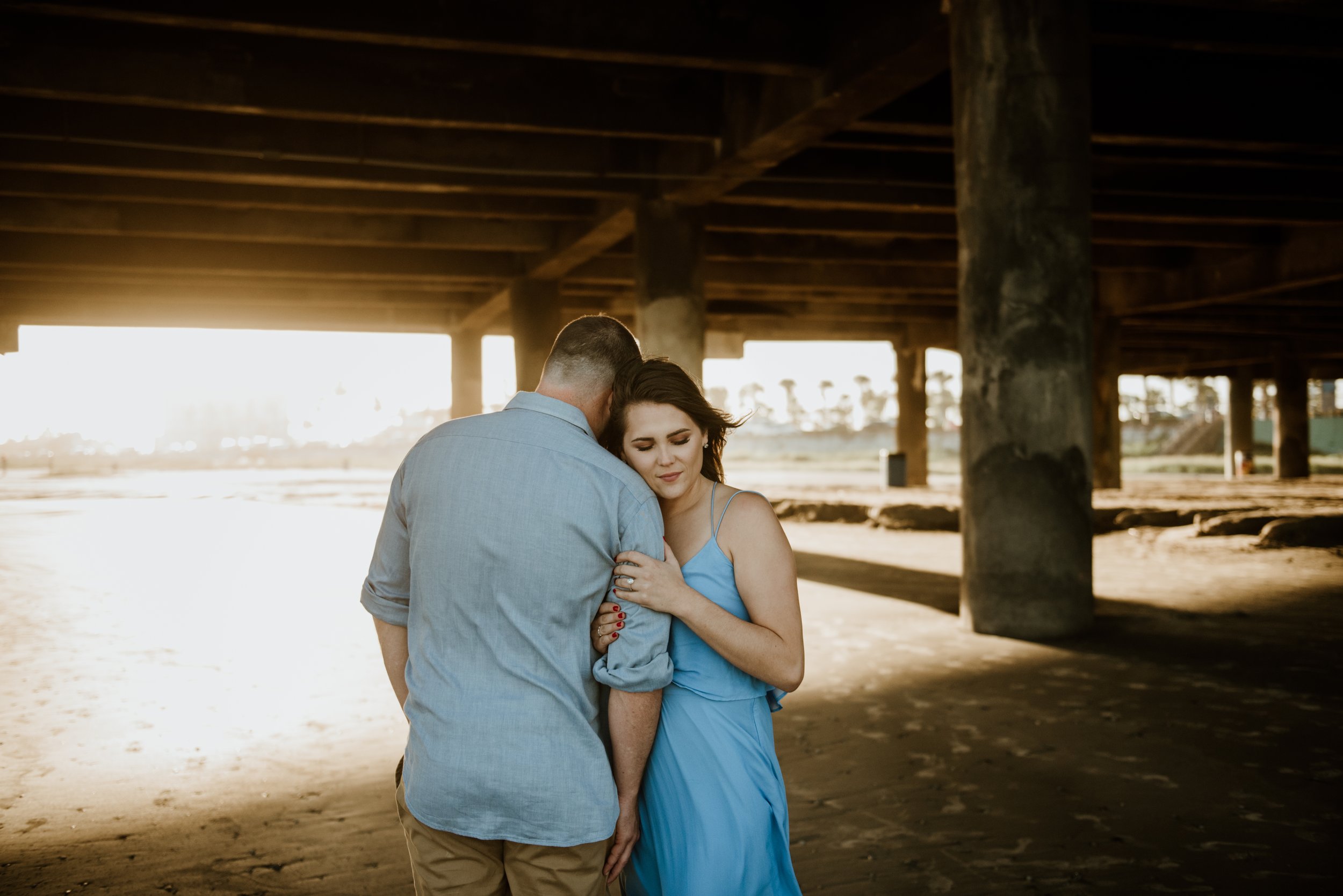A couple embracing on a beach under a pier at sunset, with the woman in a blue dress and the man in a light-colored shirt.