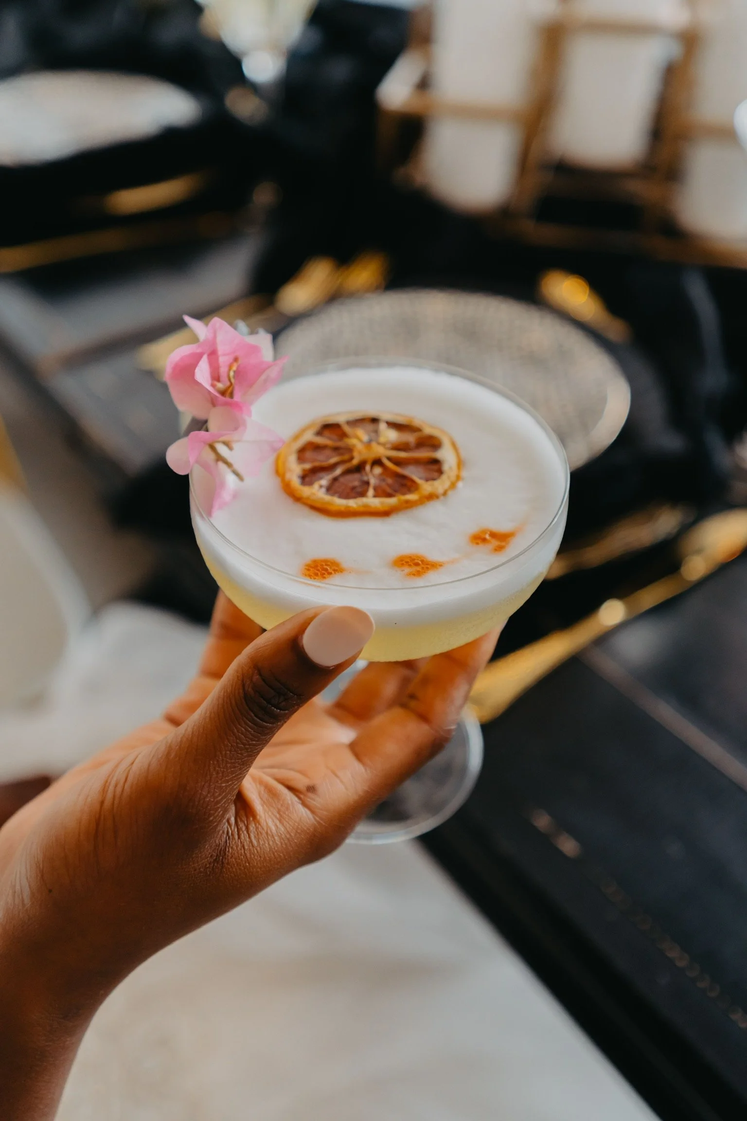 A person holding a cocktail glass topped with a slice of dried lemon and pink flowers, with a blurred background.