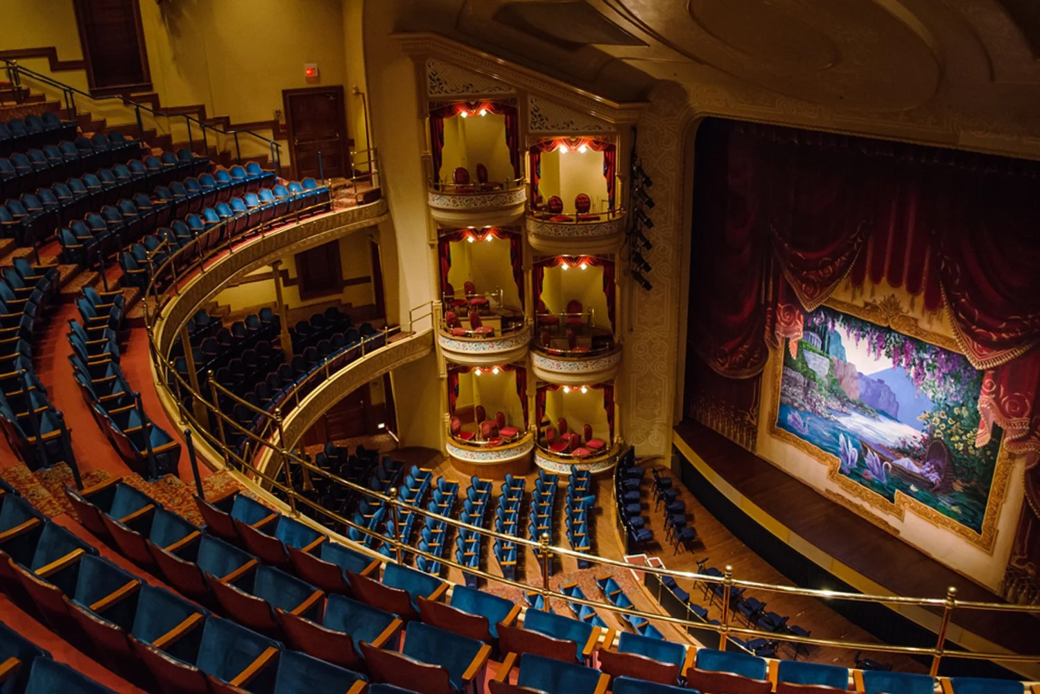 Interior view of a theater auditorium with blue seats, a stage with a large scenic backdrop, and luxurious balconies with red and gold curtains.