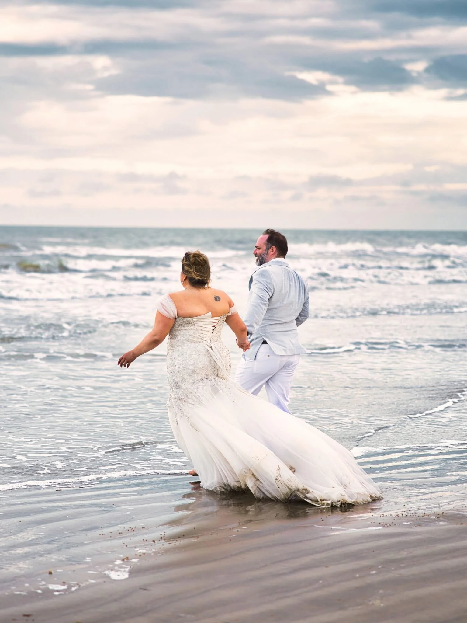 A bride and groom walking hand in hand on the beach during sunset, with waves crashing around their feet.
