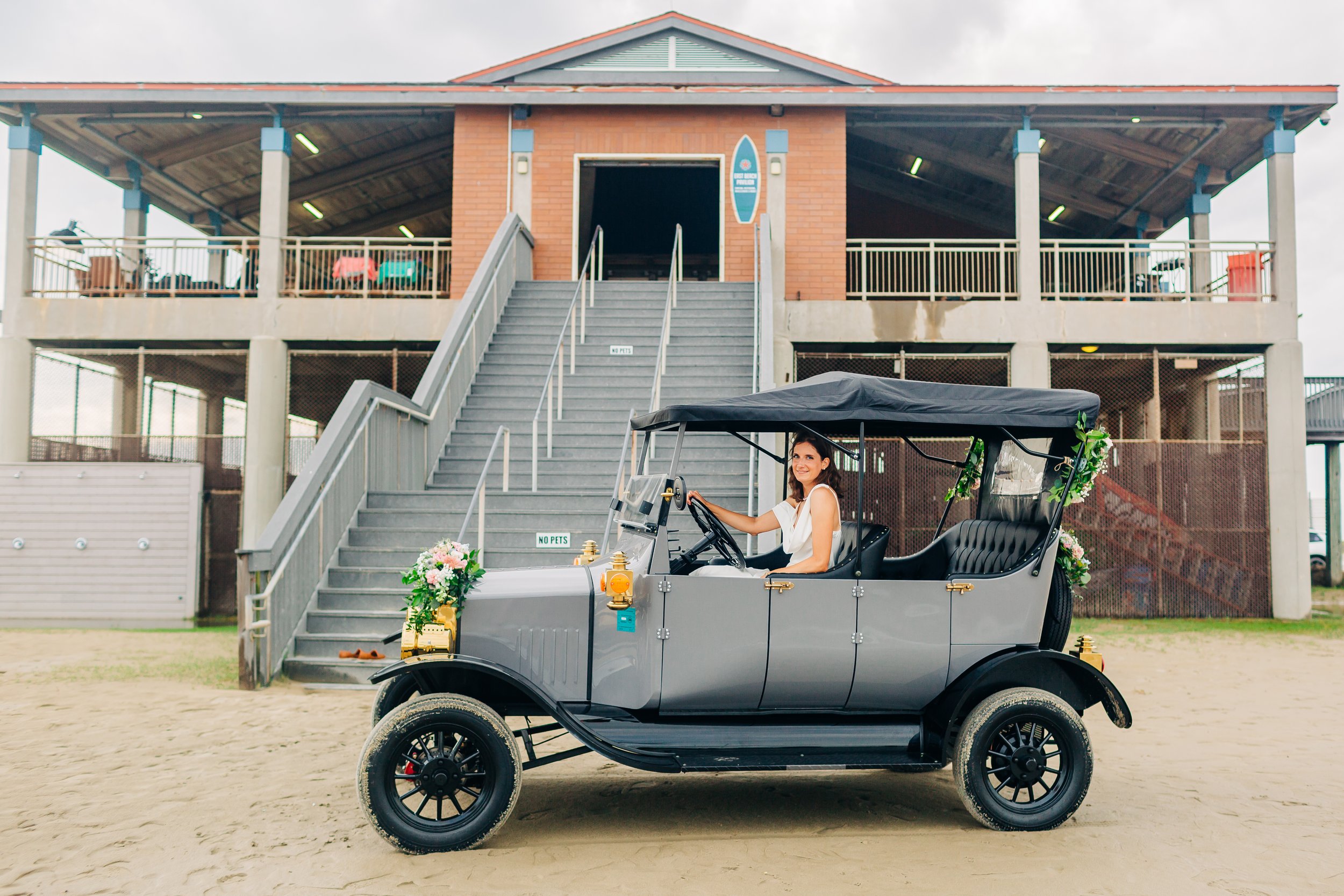 A woman dressed in white sitting in a vintage golf cart decorated with flowers, parked in front of a beachside wooden structure on a sandy path.