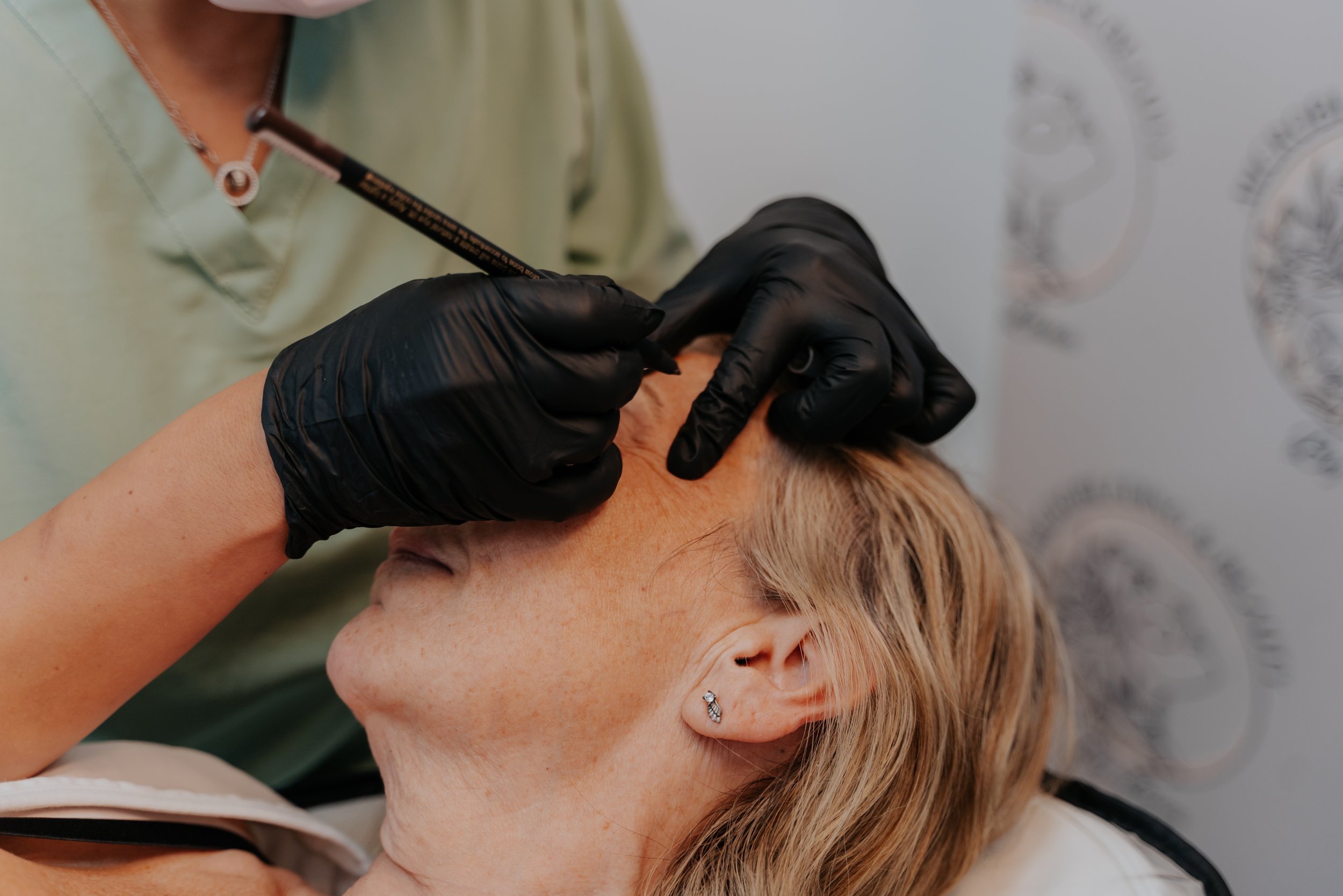 A person with blonde hair and earrings receiving a cosmetic injection or treatment around their eye area from a professional wearing black gloves.