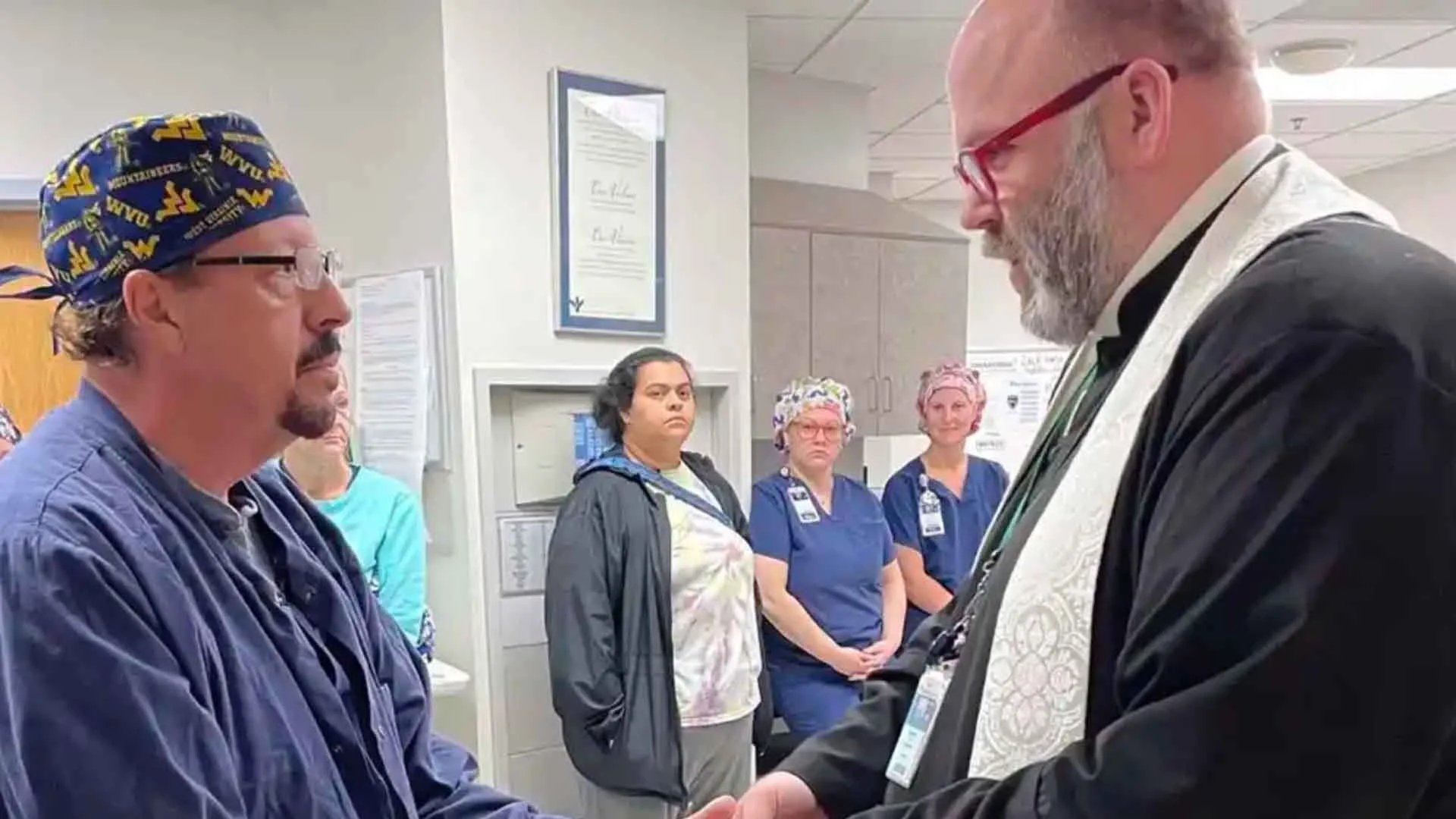 A medical chaplain holds the hands of a nurse in the foreground as his co-workers look on in the background