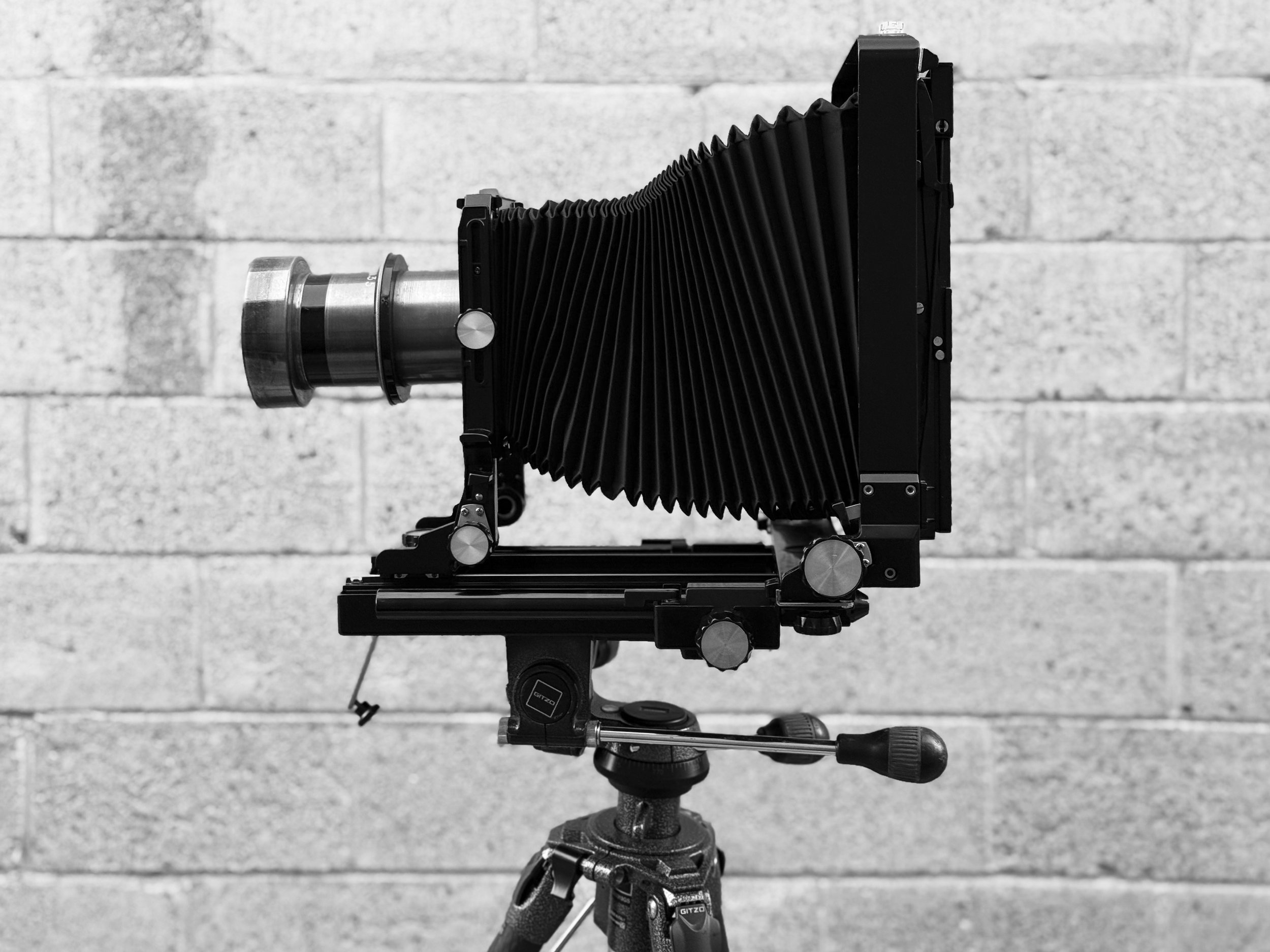 Large-format camera with bellows mounted on a tripod against a brick wall.