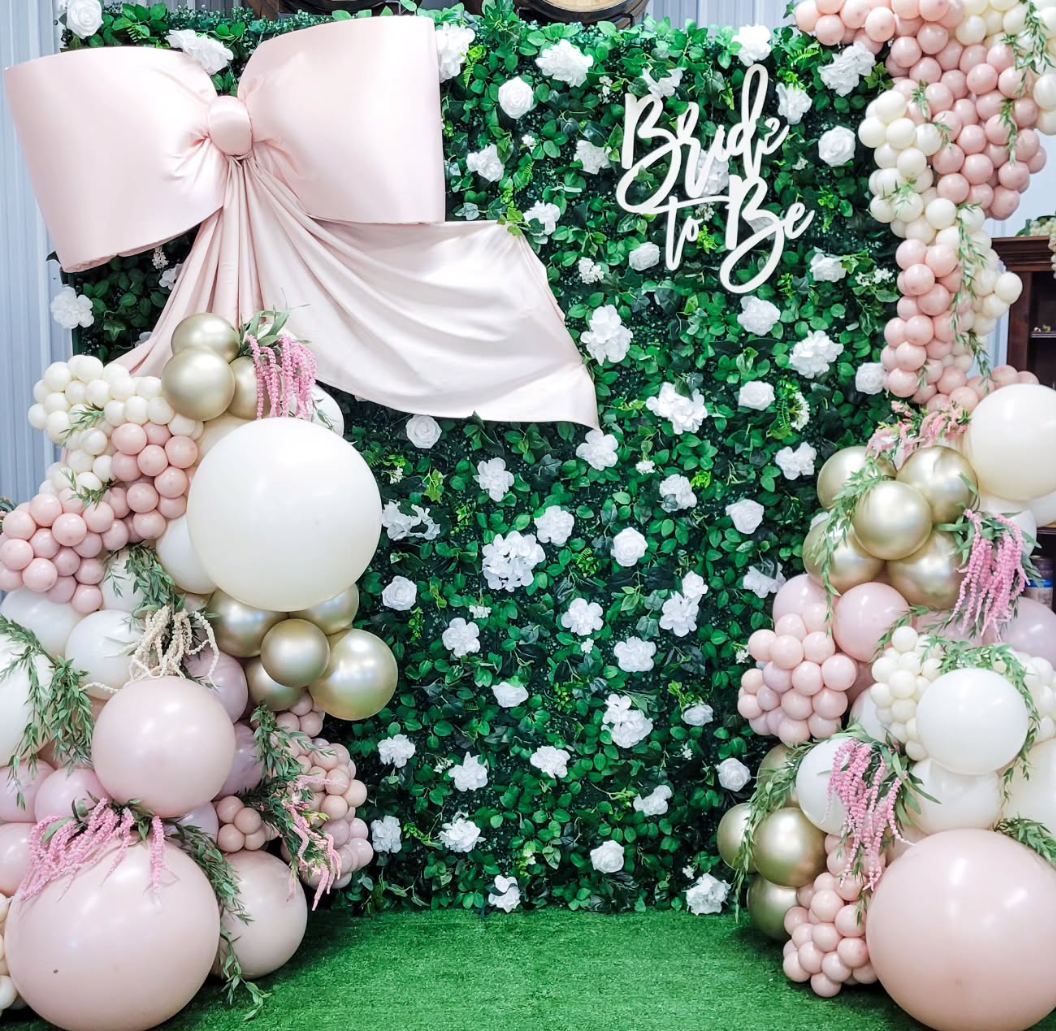 Decorative backdrop for a bridal event with green foliage, white flowers, pink and white balloons, and a pink bow with the words "Bride to Be" in white script.