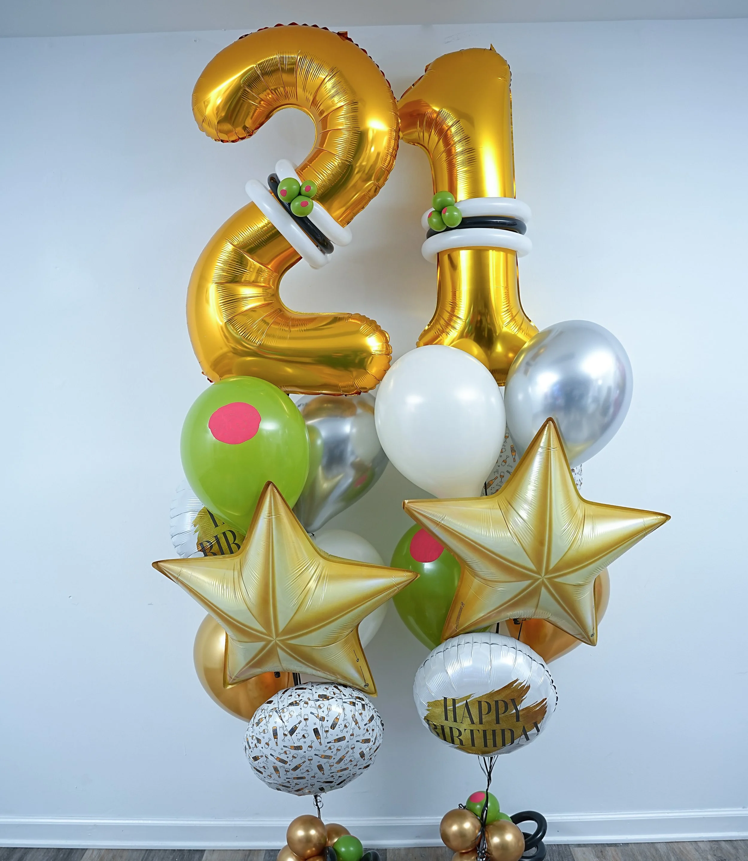 Decorative balloon arrangement for a birthday celebration featuring large gold balloons shaped as the number "21" and various smaller balloons in white, green, black, and gold, with some balloons having birthday-themed prints and messages.
