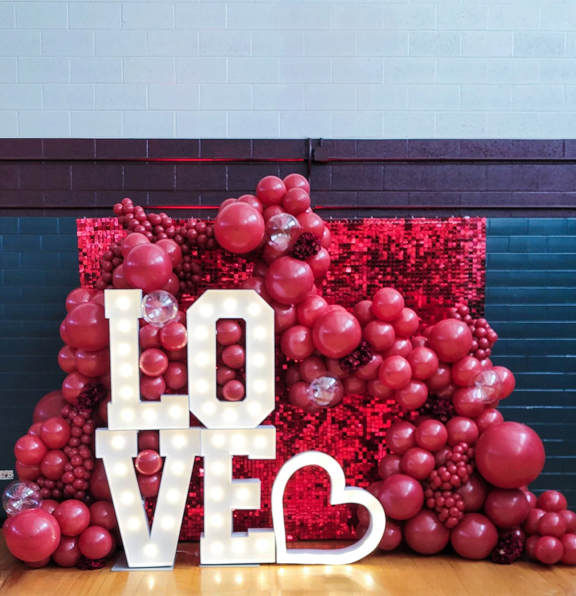 Large white illuminated letters spelling out 'LOVE' surrounded by pink balloons and a red mosaic backdrop, with a white 10 in the foreground, against a blue brick wall.