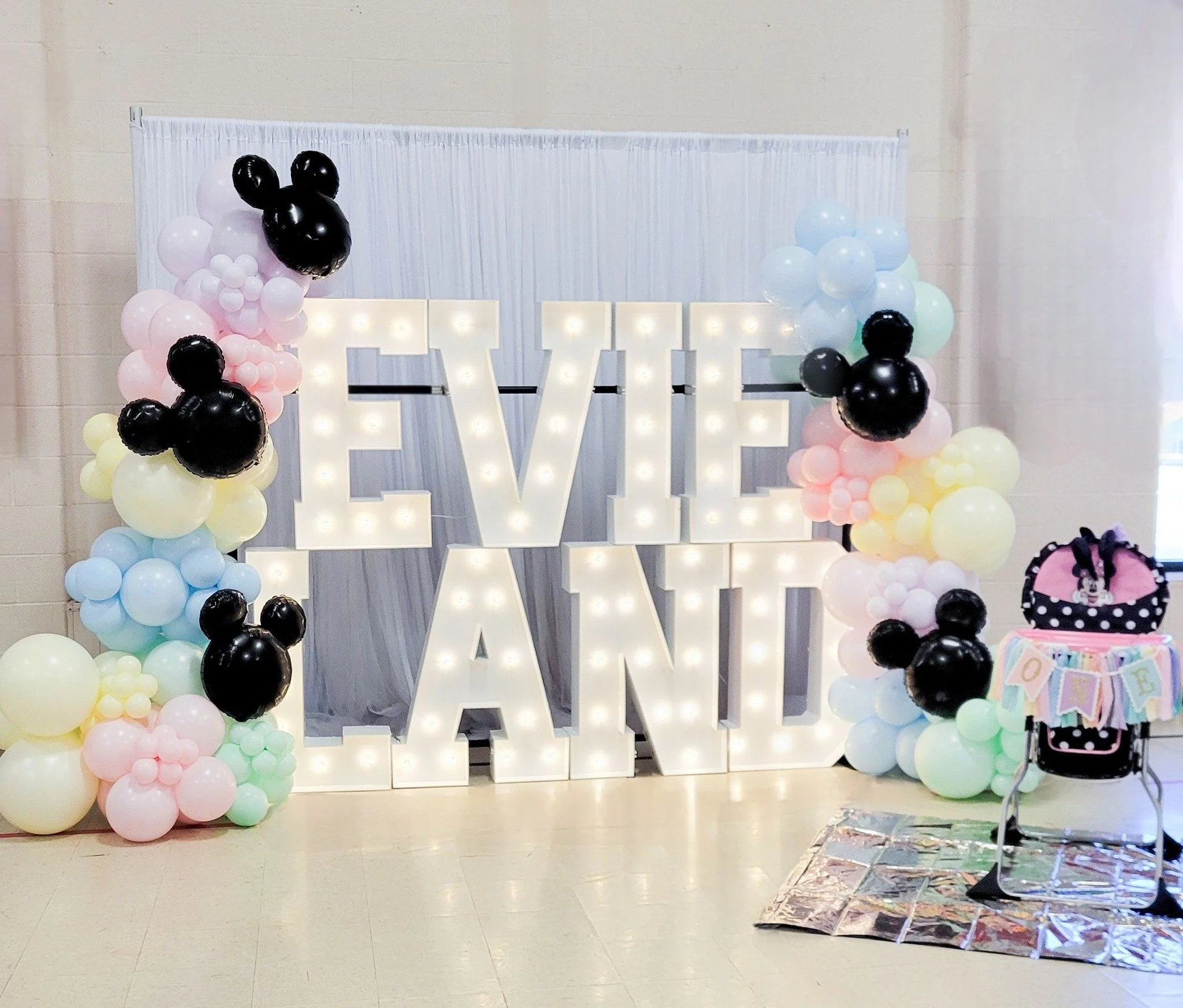 Decorative backdrop with illuminated block letters spelling 'LOVE' flanked by pastel and black balloons on the sides, likely for a celebration or party.