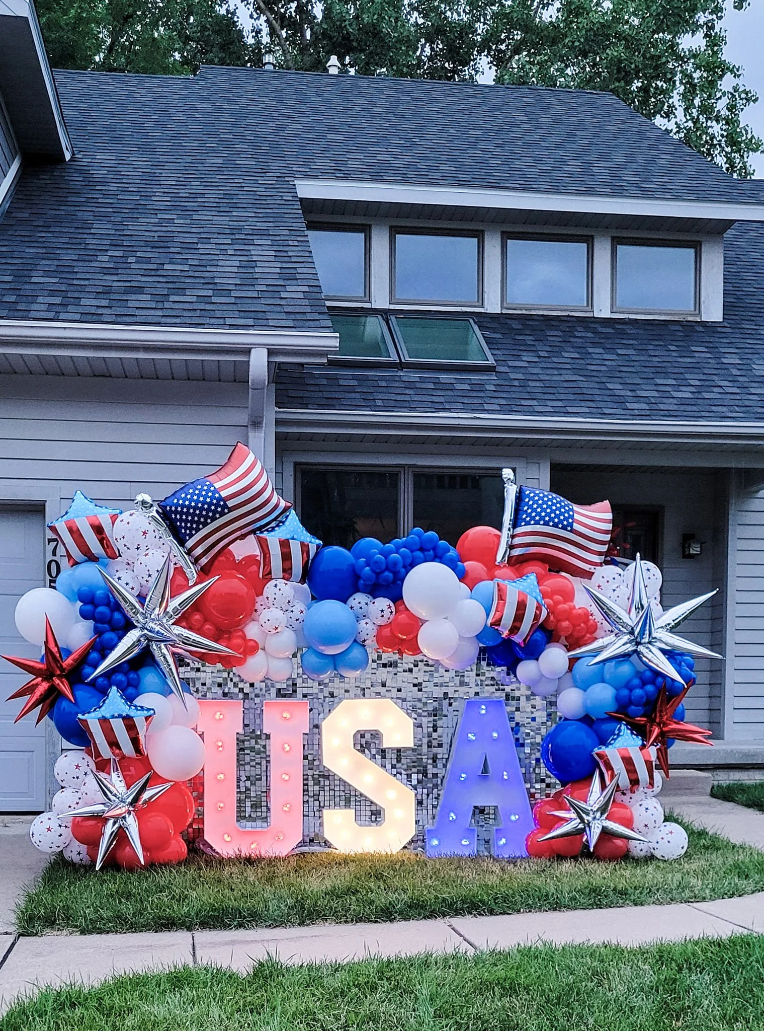 Patriotic celebration with red, white, and blue balloons, star-shaped balloons, and illuminated letters spelling out 'USA' in front of a house with a brick wall and large windows.