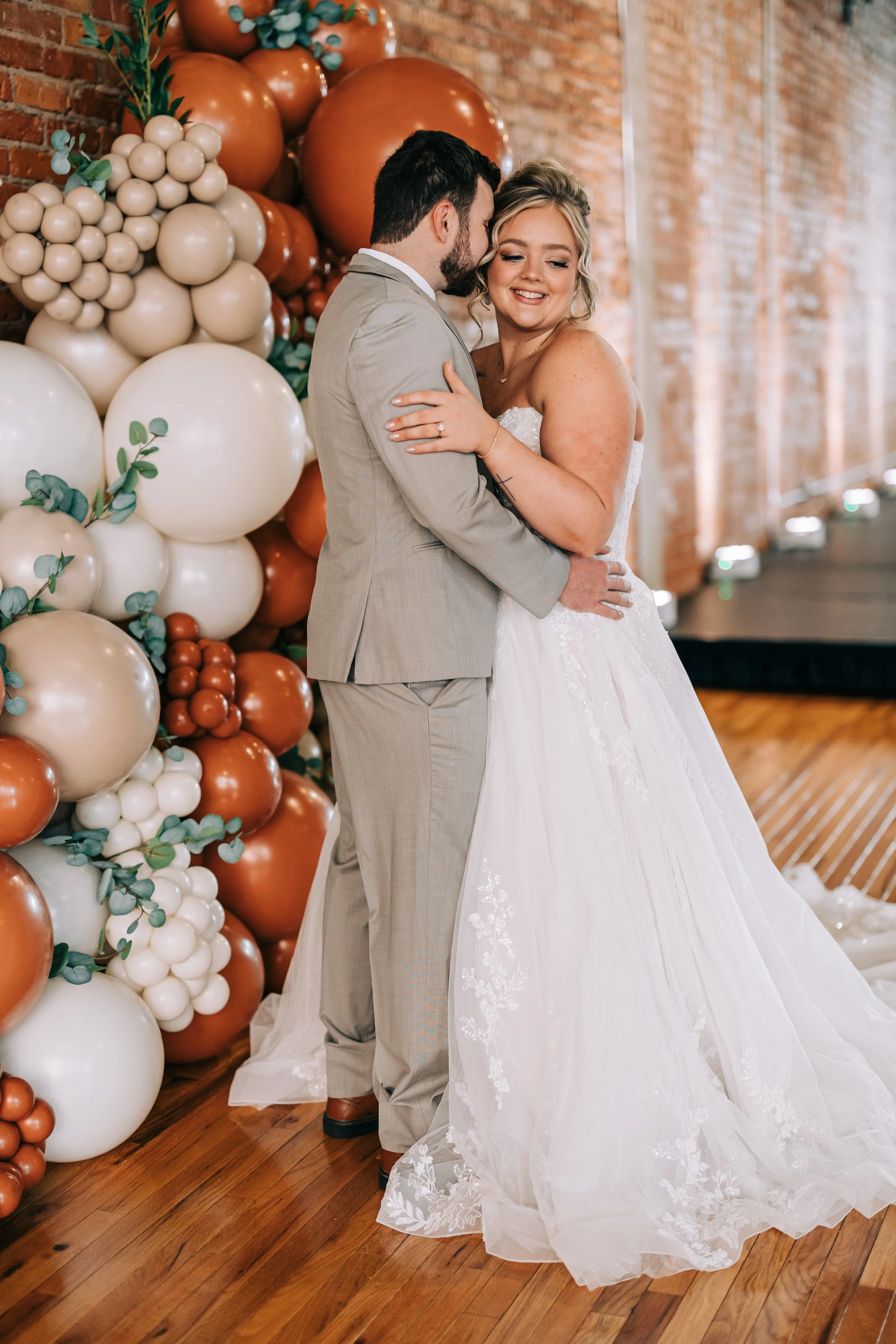 A bride and groom sharing a dance at their wedding reception, with a balloon arch decor in the background and a brick wall.