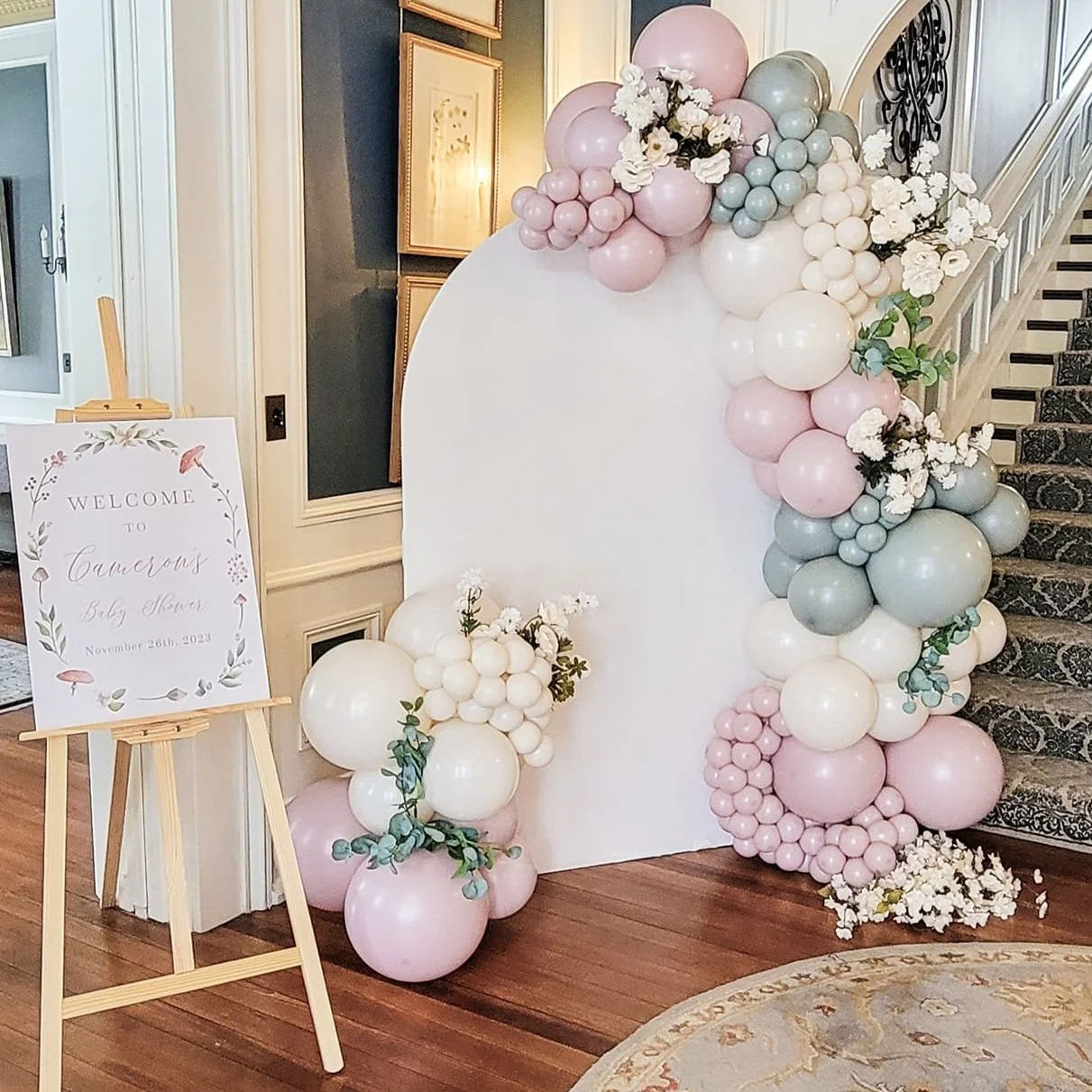 Decorative balloon arrangement with pink, gray, and white balloons, flowers, and greenery next to a staircase. A sign on an easel welcoming guests to a baby shower on November 26th, 2023, is also visible.
