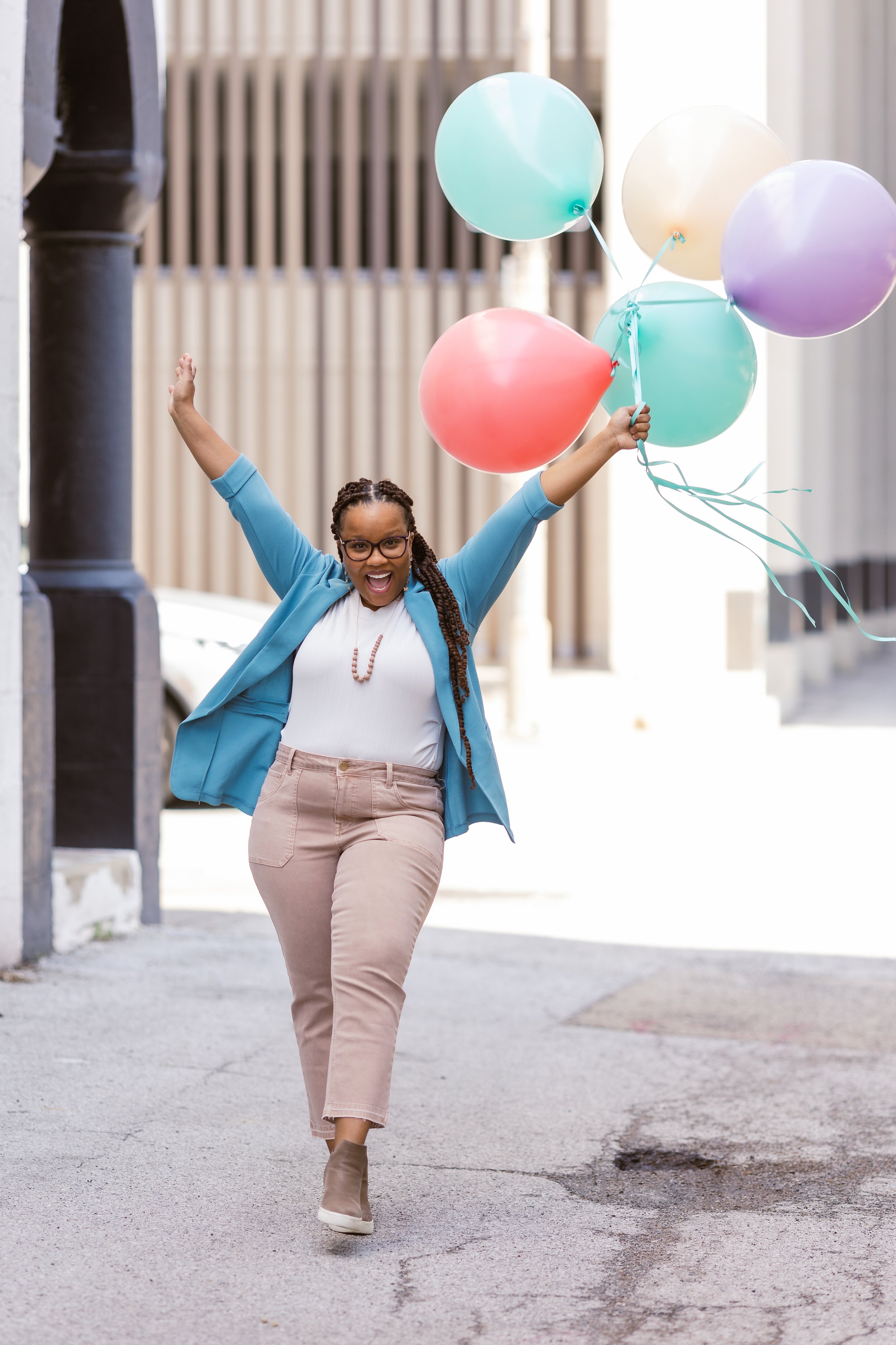 Woman walking outdoors holding colorful balloons, smiling and raising her arms in celebration.