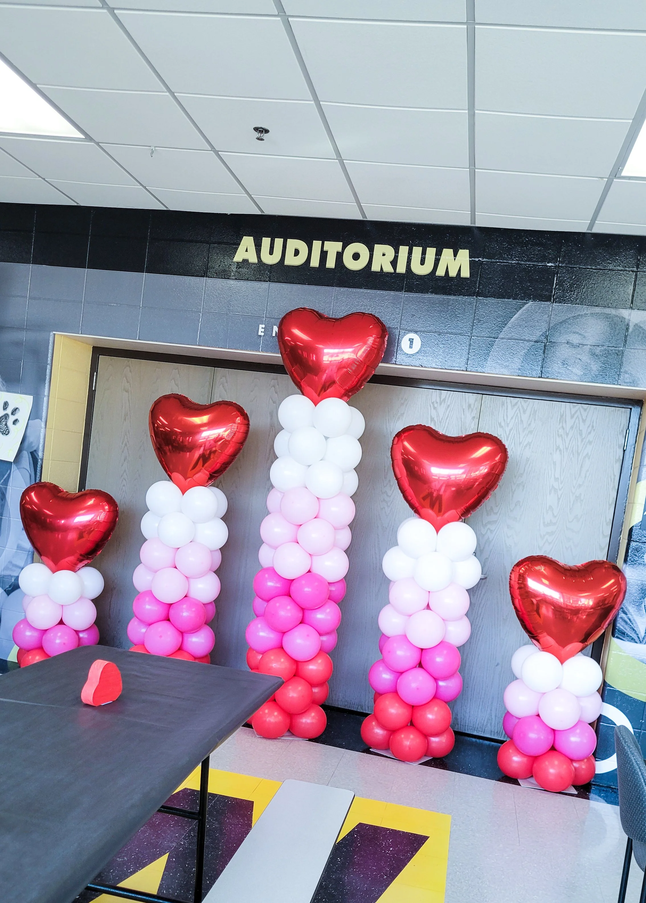 Decorative columns made of pink, white, and red balloons with large red heart-shaped balloons on top, placed in front of a sign that reads 'Auditorium' in a school or event space.