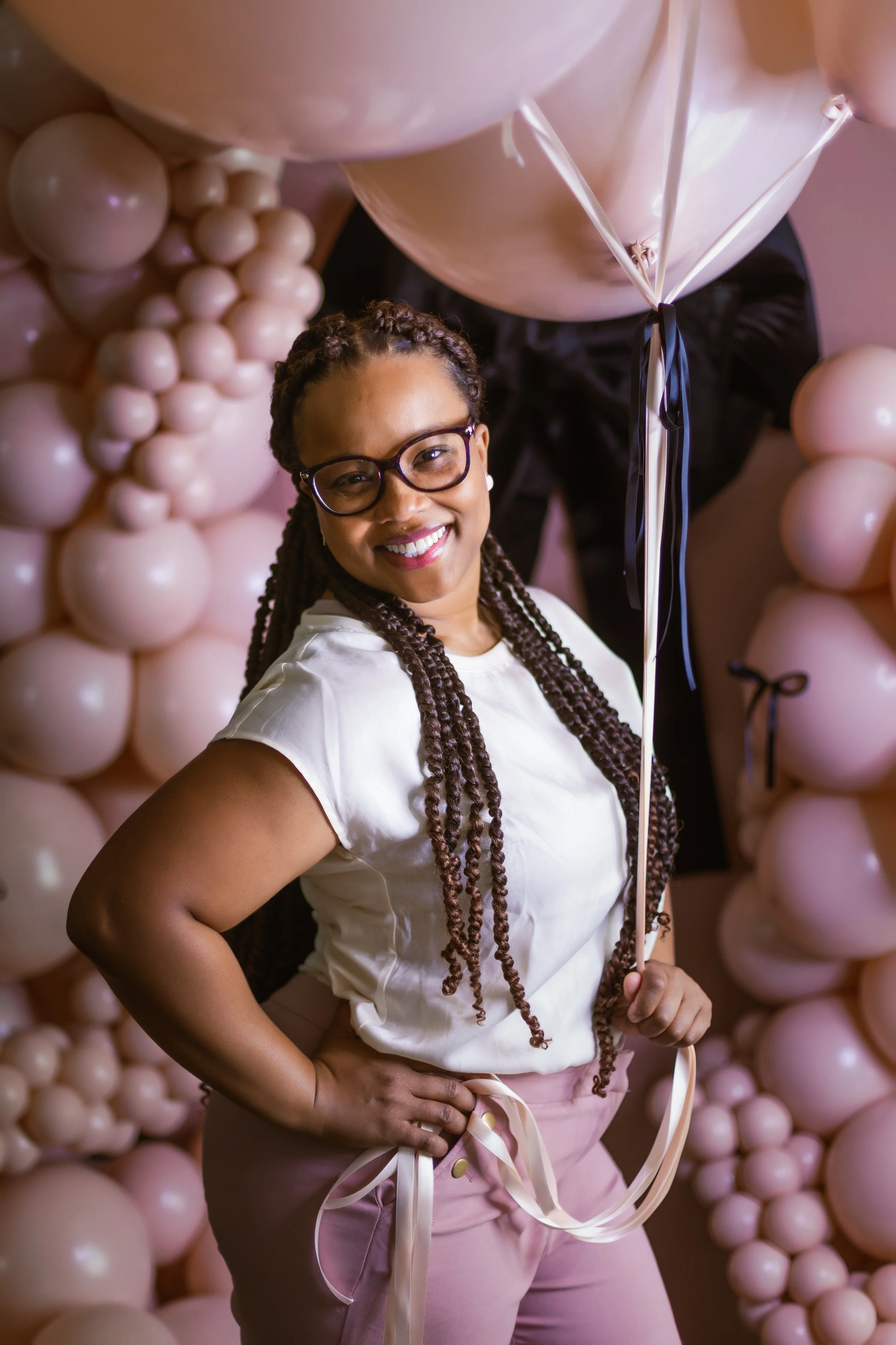 A woman with glasses and braided hair smiling, holding a pink balloon, standing in front of a backdrop of pink balloons.