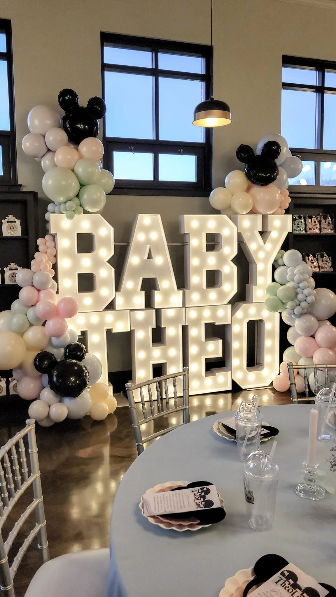 A celebration setup with illuminated large letters spelling 'BABY' and balloons in pastel colors and black, pink, white, green. Chairs and a table with drinking glasses are in the foreground.