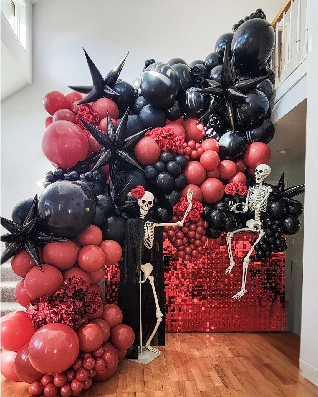 Halloween decoration featuring a large balloon arch with red and black balloons, black star-shaped balloons, skeleton figures, red roses, and a shiny red and black backdrop.