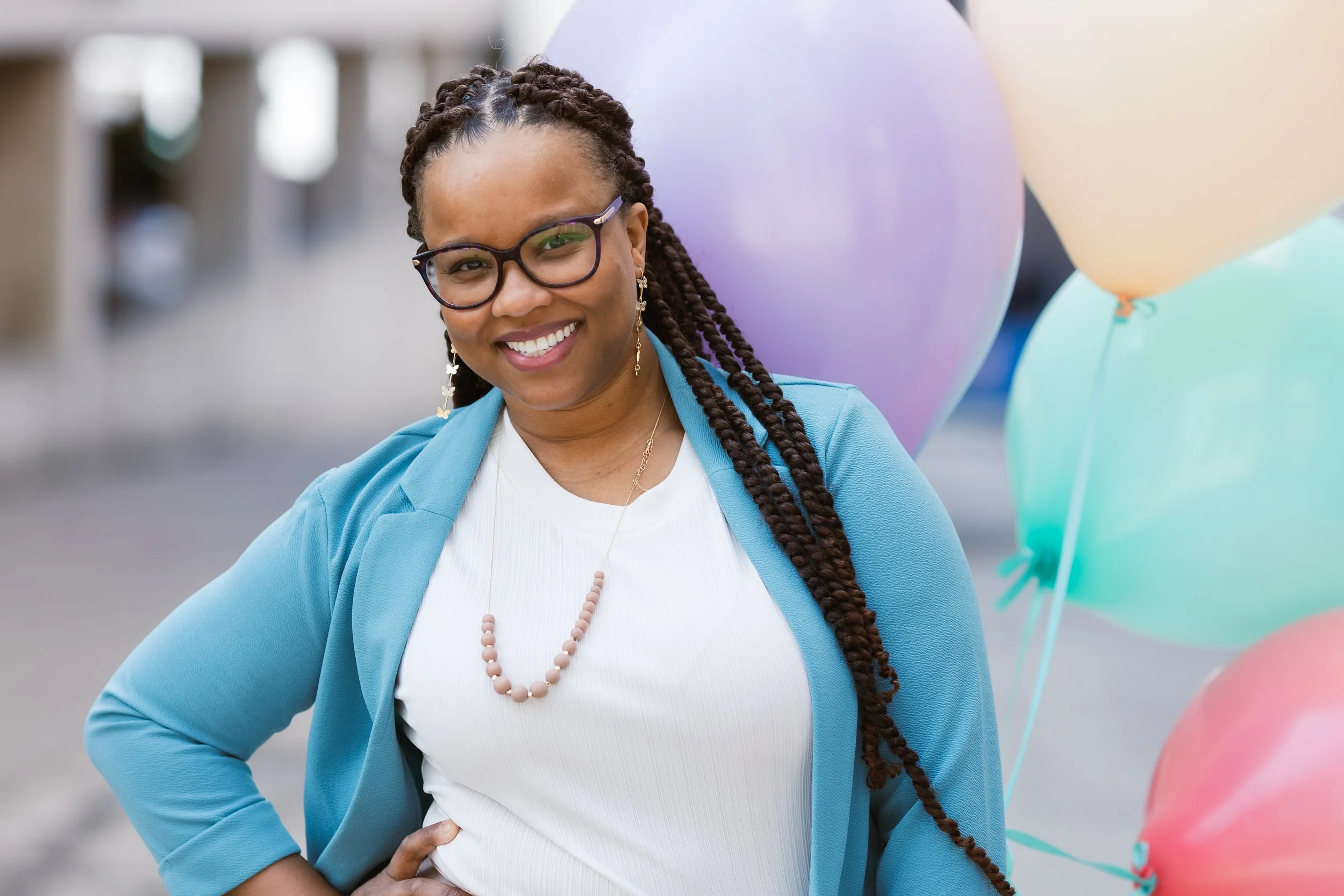 Smiling African American woman wearing glasses and a turquoise blazer, holding colorful balloons, with her hand on her hip, outdoors.