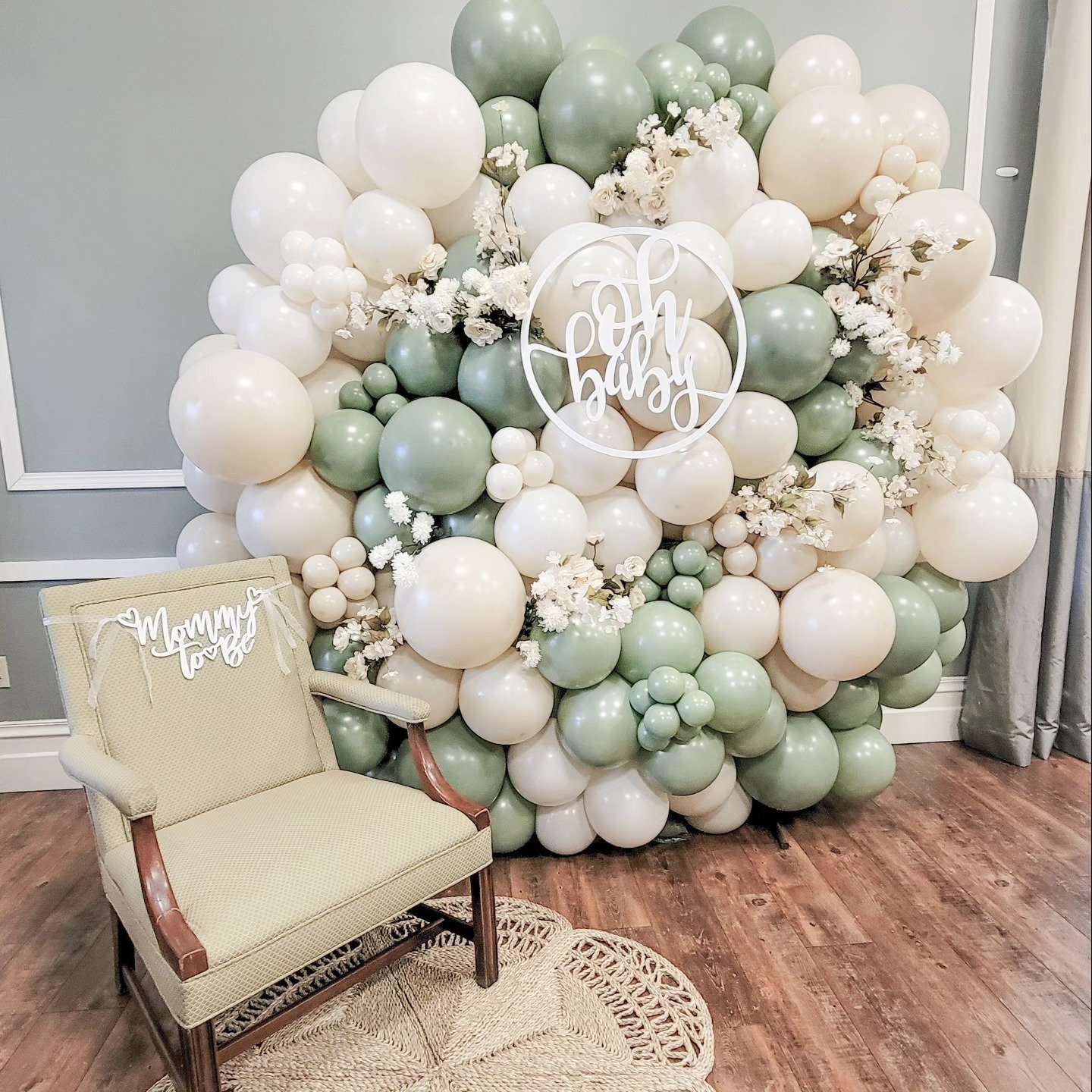 A large balloon arrangement in soft white, cream, and sage green colors, decorated with white flowers, with a circular sign that says 'Oh Baby' in cursive. There is a cream-colored vintage chair with a 'Mummy Love' sign attached to the back, sitting on a round woven rug in front of the balloon display.