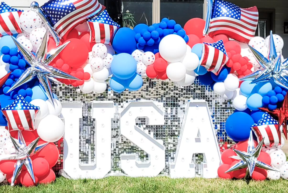 Decorative display with patriotic balloons in red, white, and blue, American flags, and large illuminated letters spelling "USA," likely for a Fourth of July celebration.