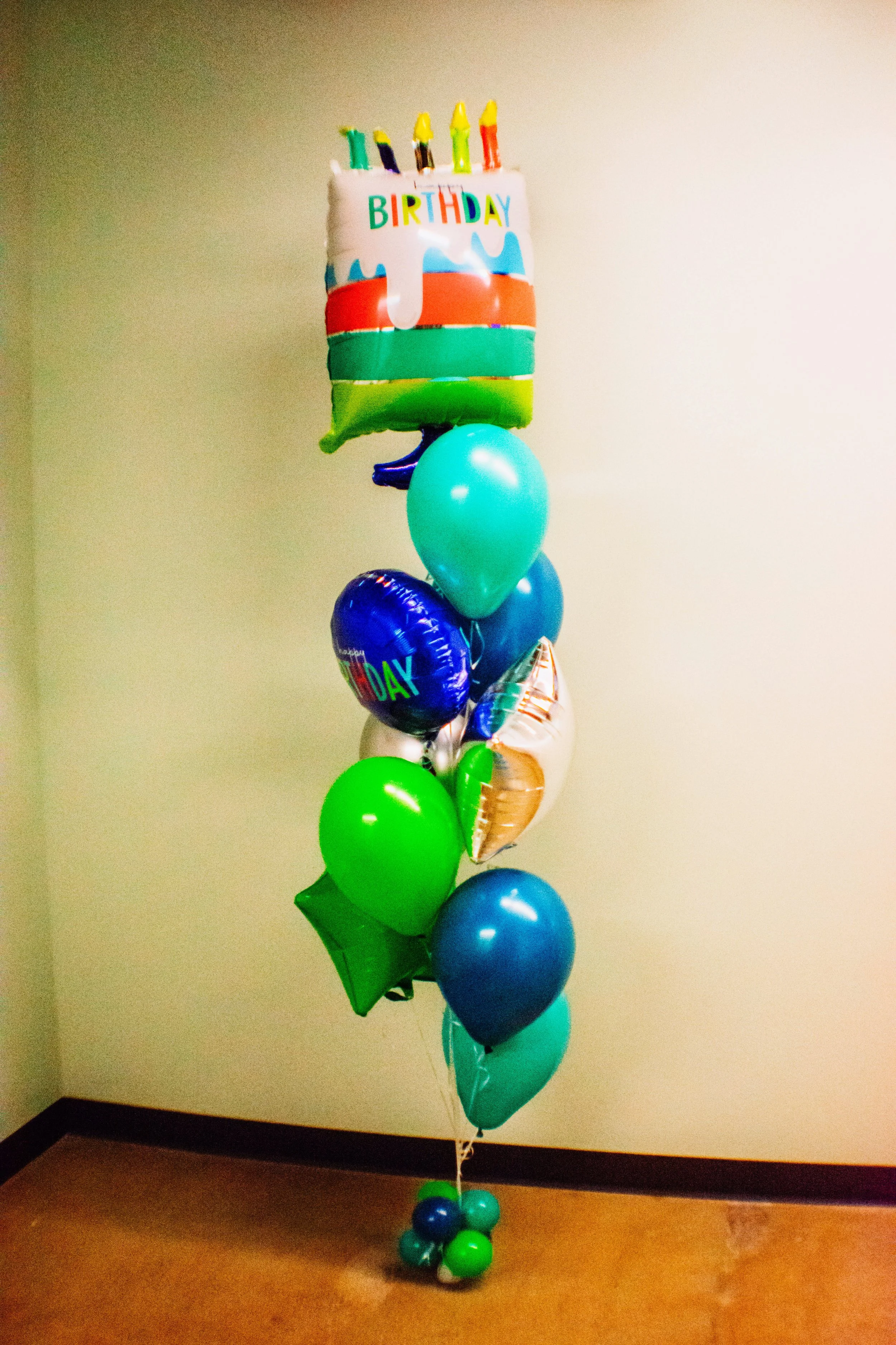 A colorful birthday balloon arrangement with a large rectangular balloon at the top that has a birthday cake design with candles and the word 'BIRTHDAY' on it. Below it are various balloons in shades of blue, green, and metallic, tied together and arranged in a column, standing against a plain wall in a room with brown flooring.
